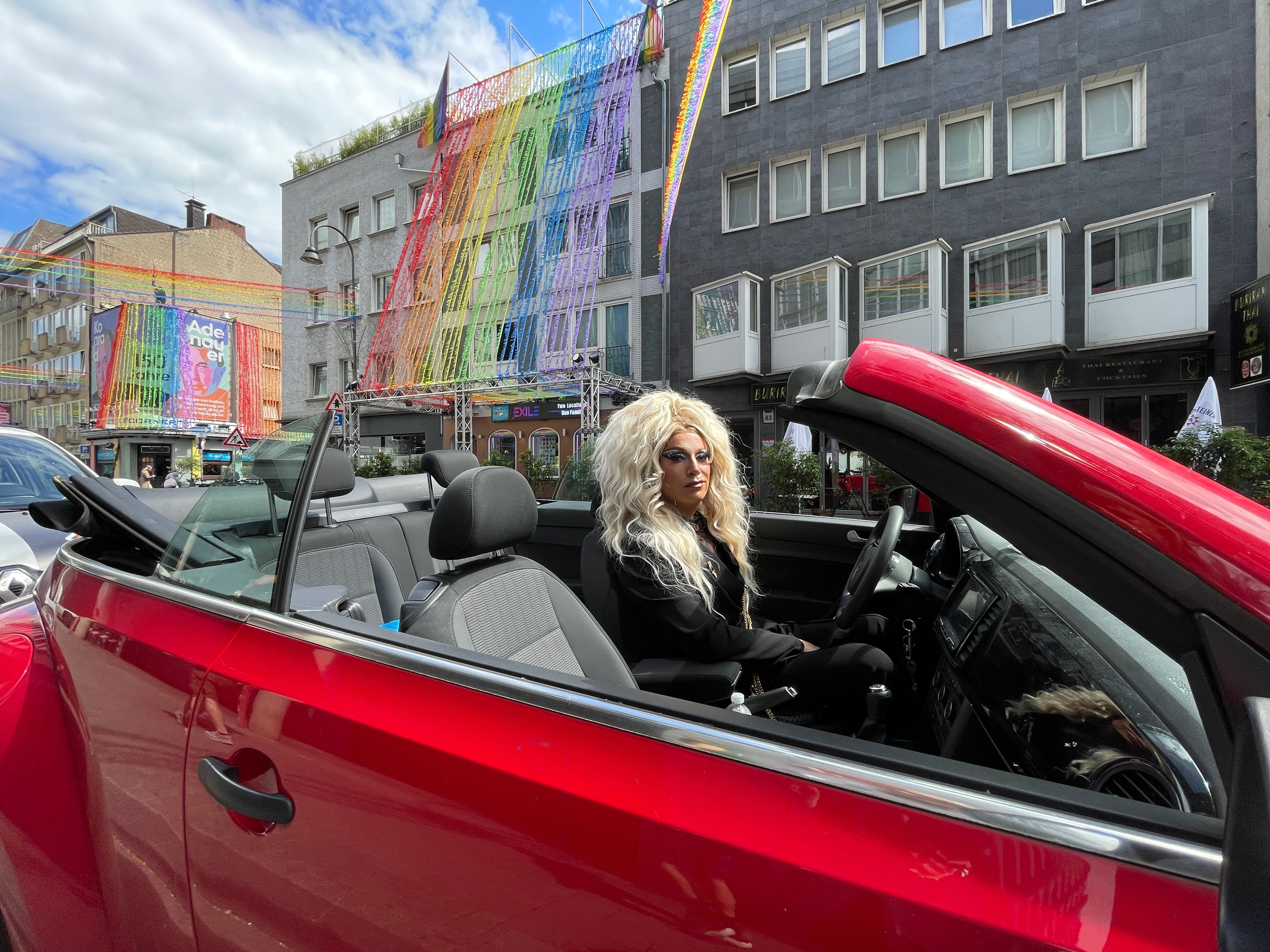 Ein als Frau mit langem blonden Haar verkleideter Mann sitzt in einem roten Cabrio. Im Hintergrund sind Fassaden mit Regenbogen-Deko zu sehen.
