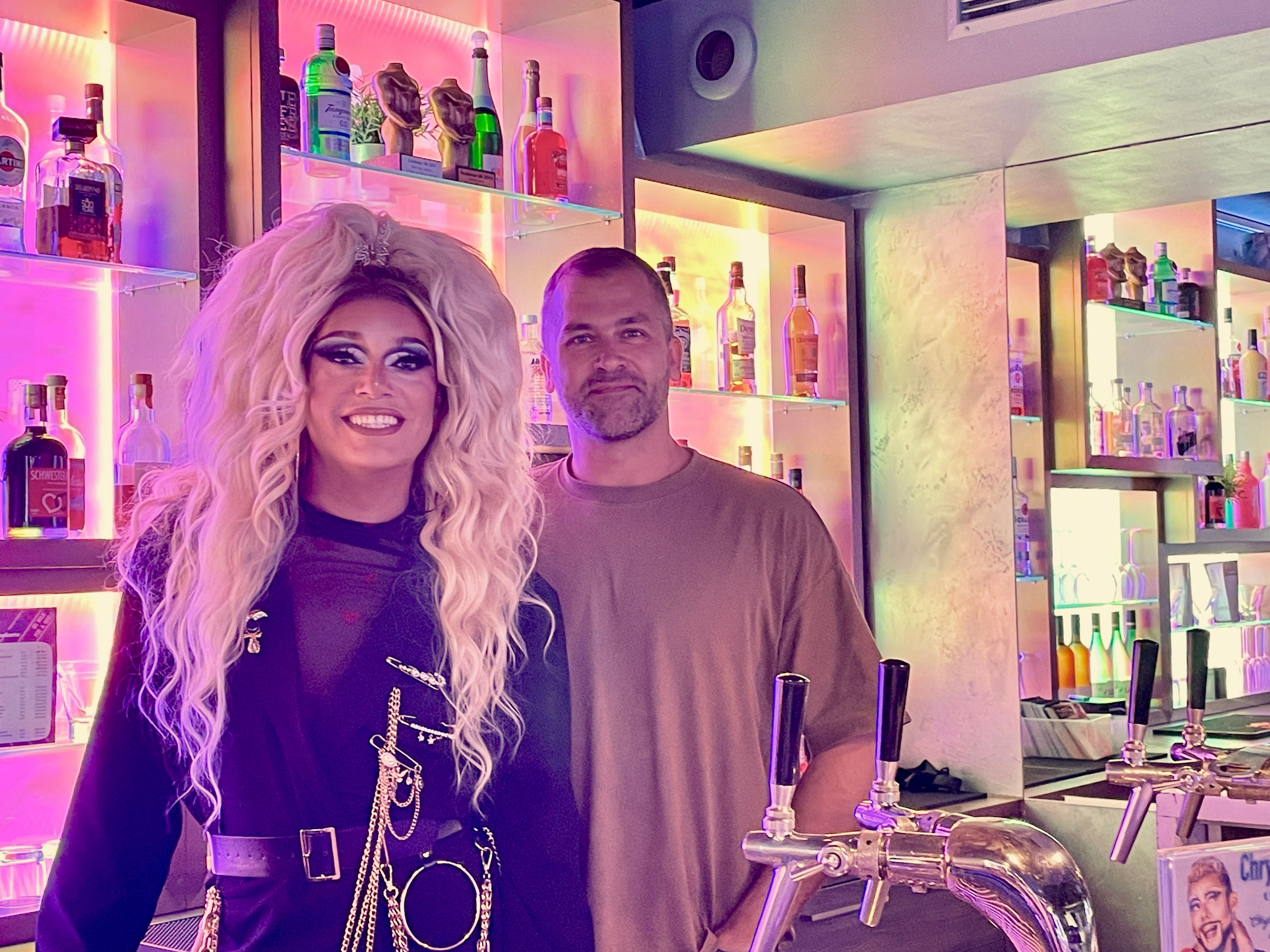 Eine Drag Queen mit langer blonder Perücke steht hinter dem Tresen einer Bar neben einem Mann im braunen Pulli mit Dreitagebart.