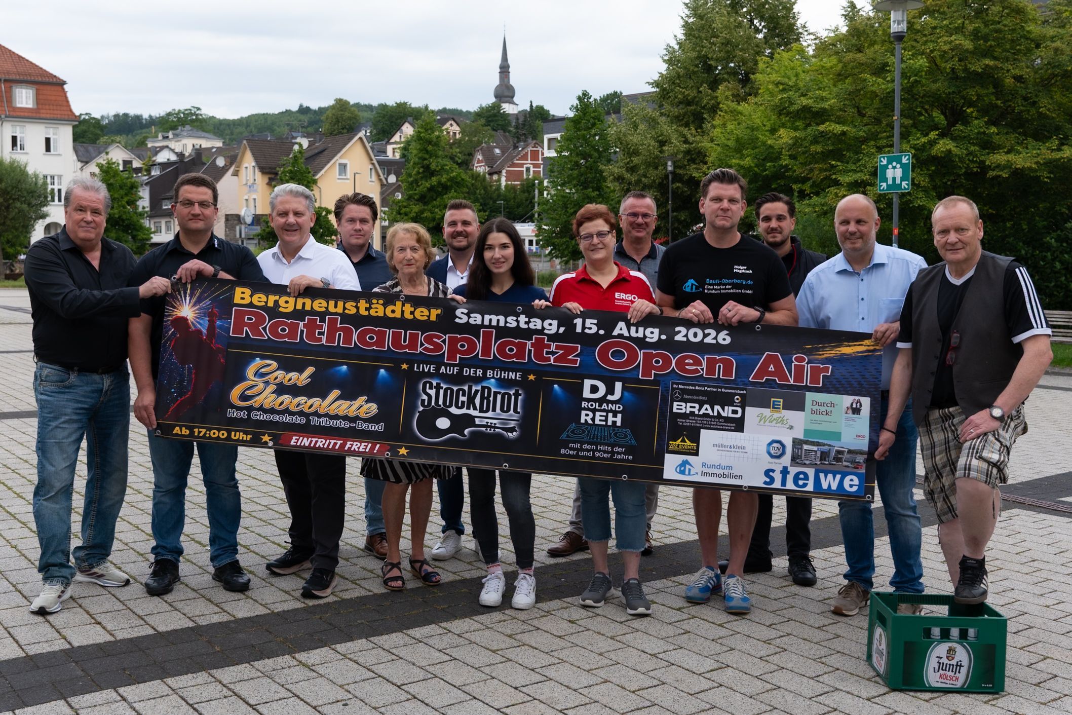 Bei einem Gruppenfoto halten mehrere Menschen ein Banner, auf dem Werbung für das Rathausplatz-Open-Air in Bergneustadt am 15. August zu lesen ist.