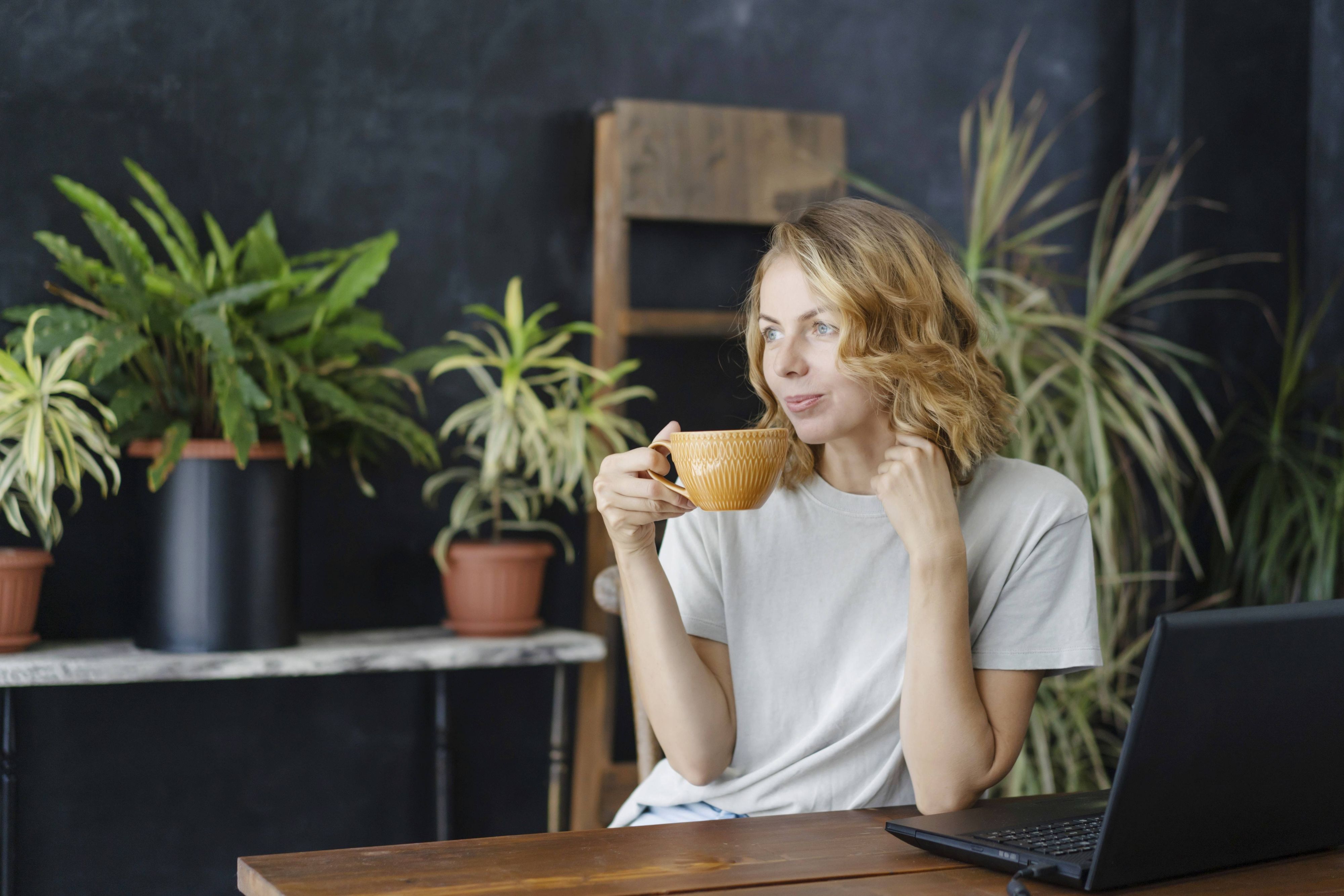 Eine Frau trinkt Kaffee und genießt an ihrem Arbeitstisch mit einem Laptop in einem gemütlichen Heimbüro mit tropischen Pflanzen im Hintergrund.