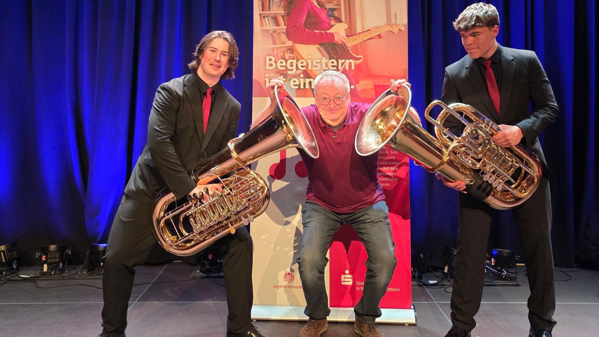 Zwei junge Tuba-Spieler und ein alter Mann stehen vor einem Plakat.