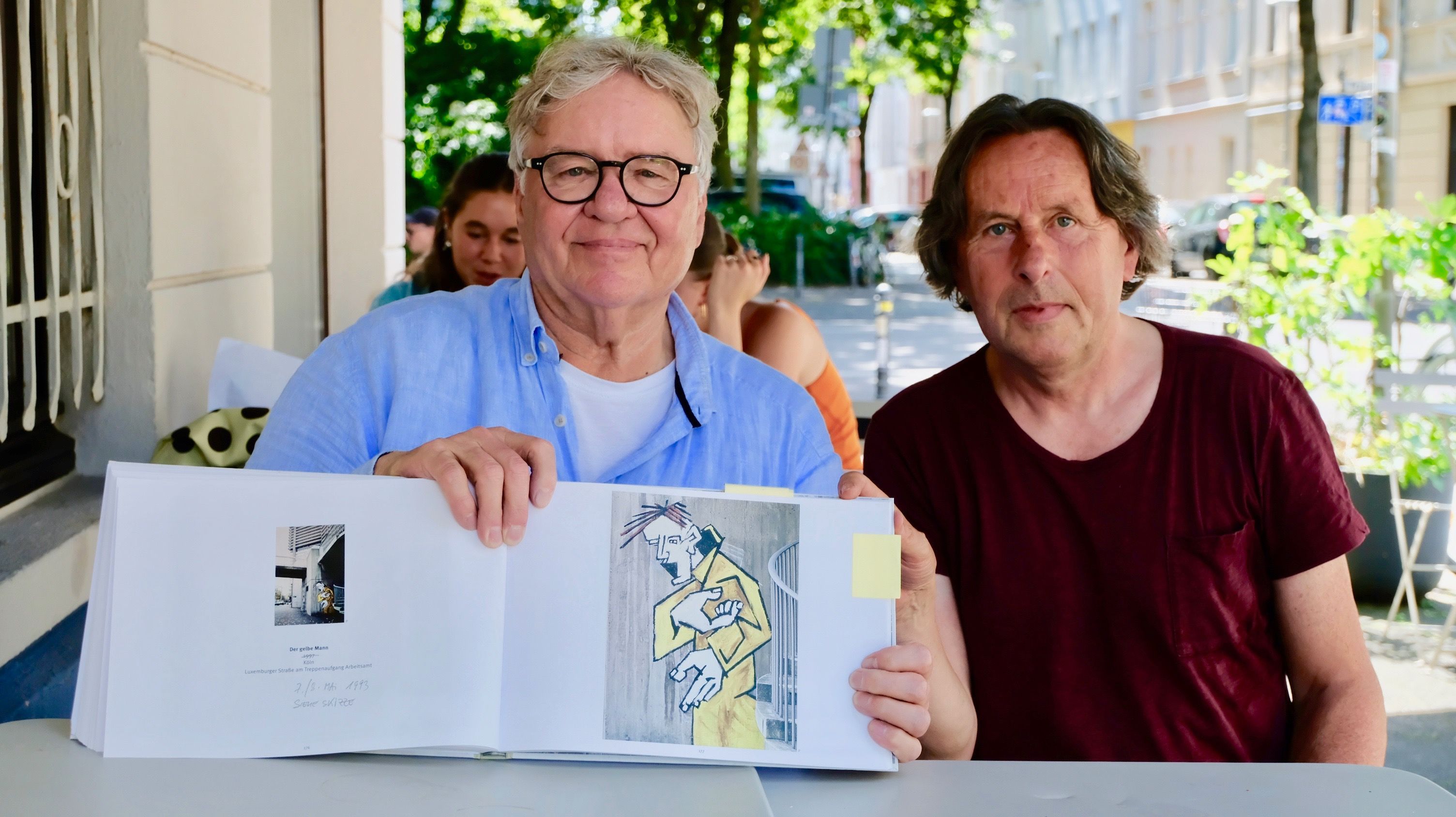 Ein Mann mit Brille sitzt neben einem Mann mit Prinz-Eisenherz-Frisur im weinroten T-Shirt. Der Mann mit Brille stützt einen aufgeklappten Bildband auf einem Tisch auf.