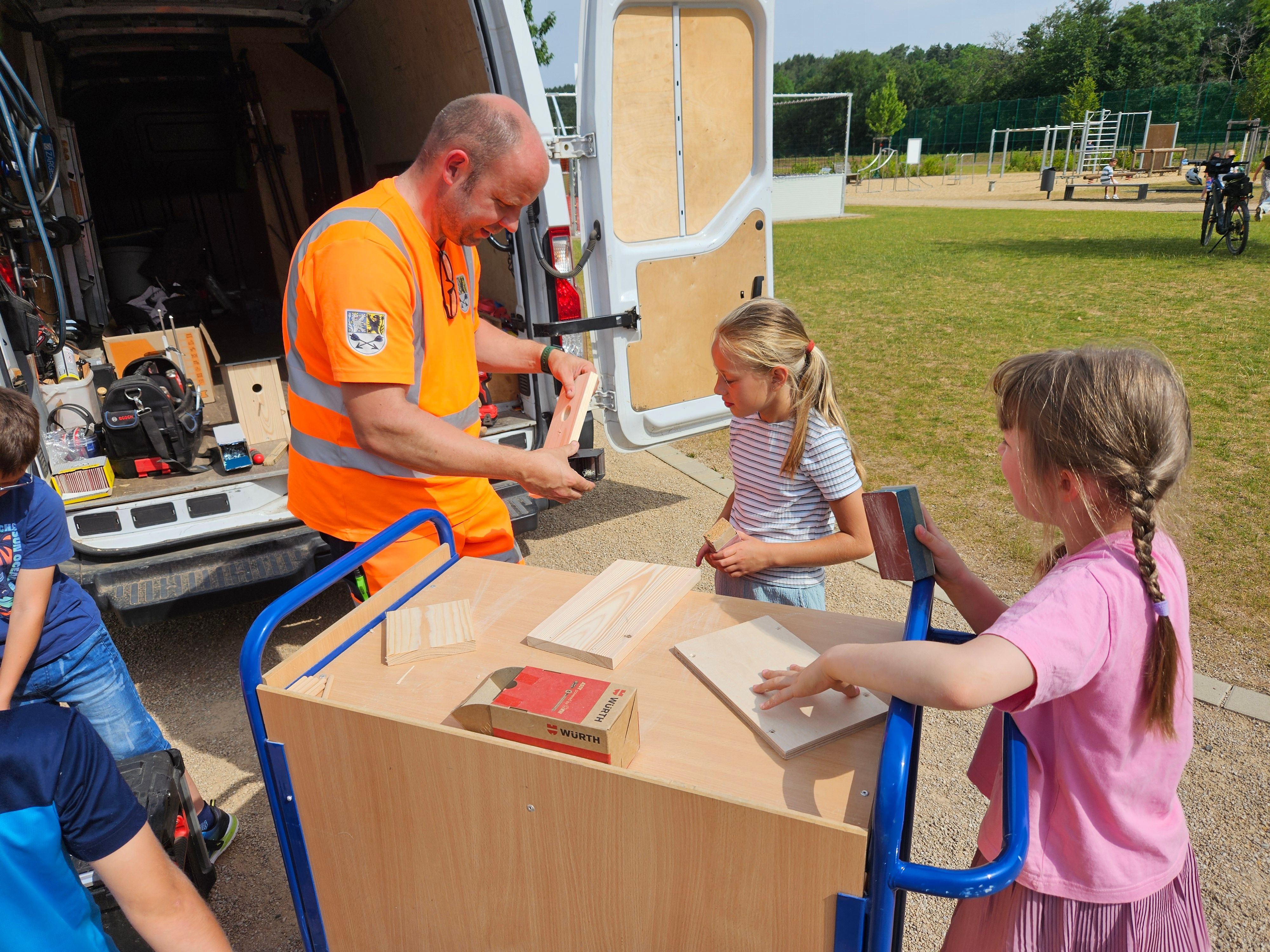 Ein Mitarbeiter des Bauhofs und zwei Mädchen arbeiten mit Holzteilen.