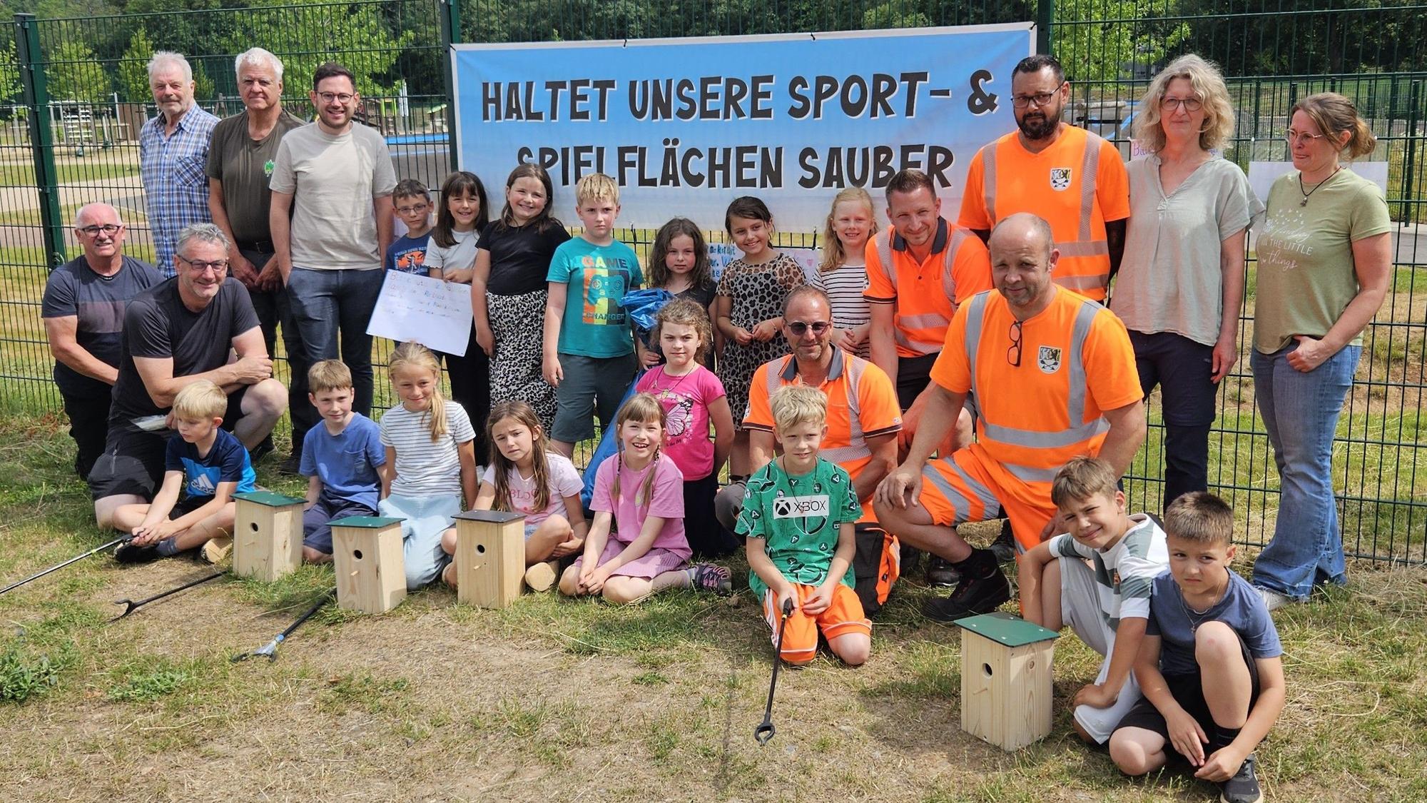 Die Helfer und die Kinder stehen und knien vor einem Banner mit der Aufschrift „Haltet unsere Sport- und Spielflächen sauber“.