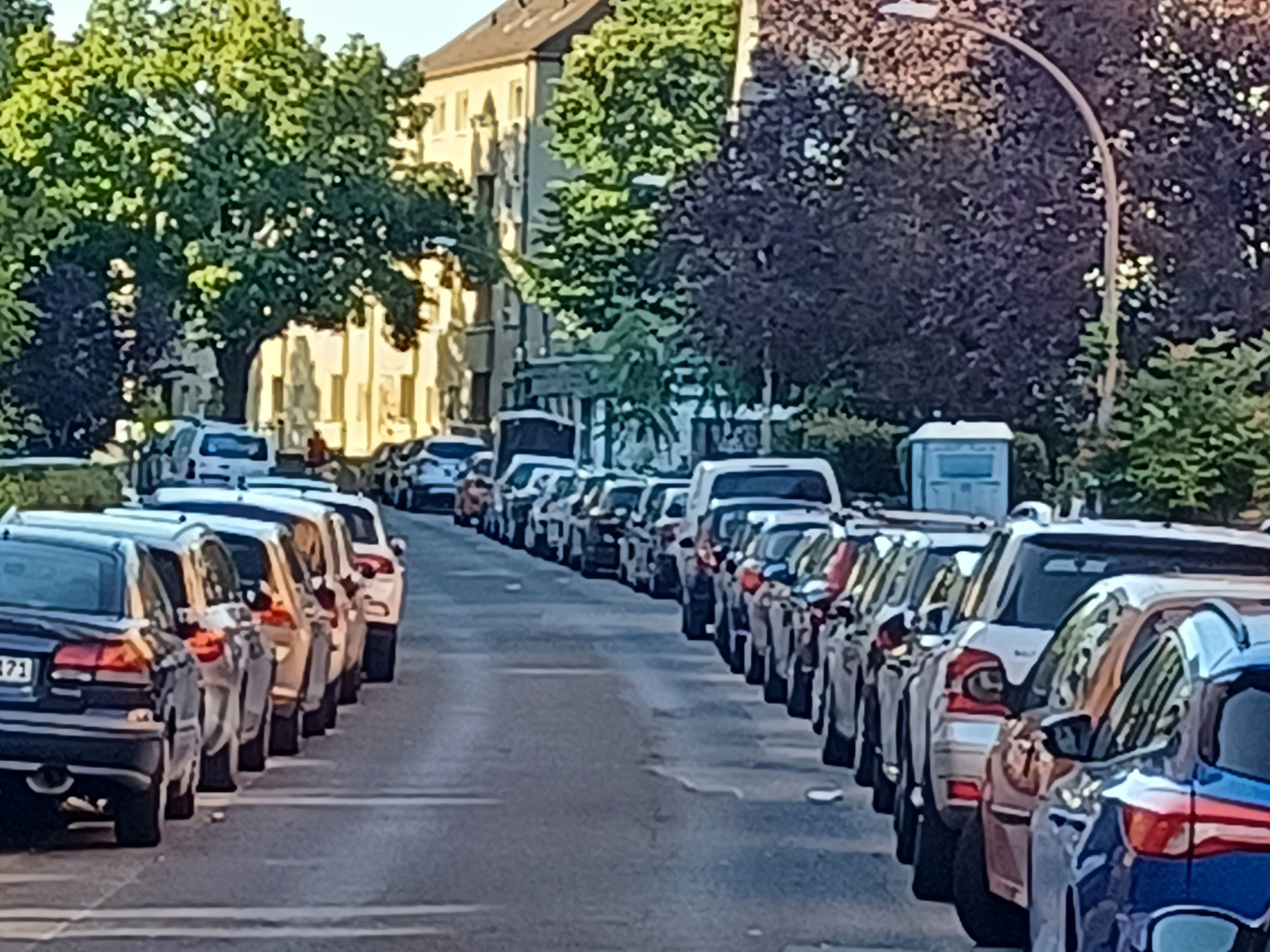 An einer mit Bäumen bestandenen Straße parken links und rechts Autos.