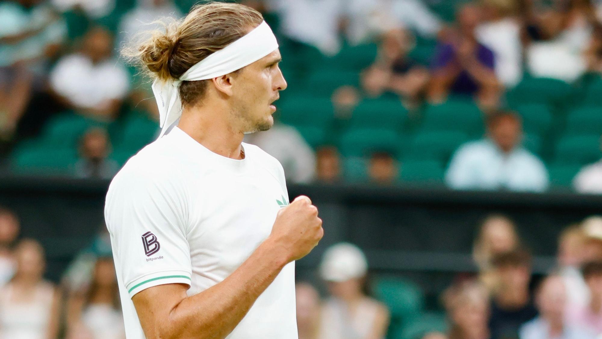 Alexander Zverev steht auf dem Tennisplatz und zeigt die Faust. Er hat ein weißes Shirt und ein weißes Stirnband an.