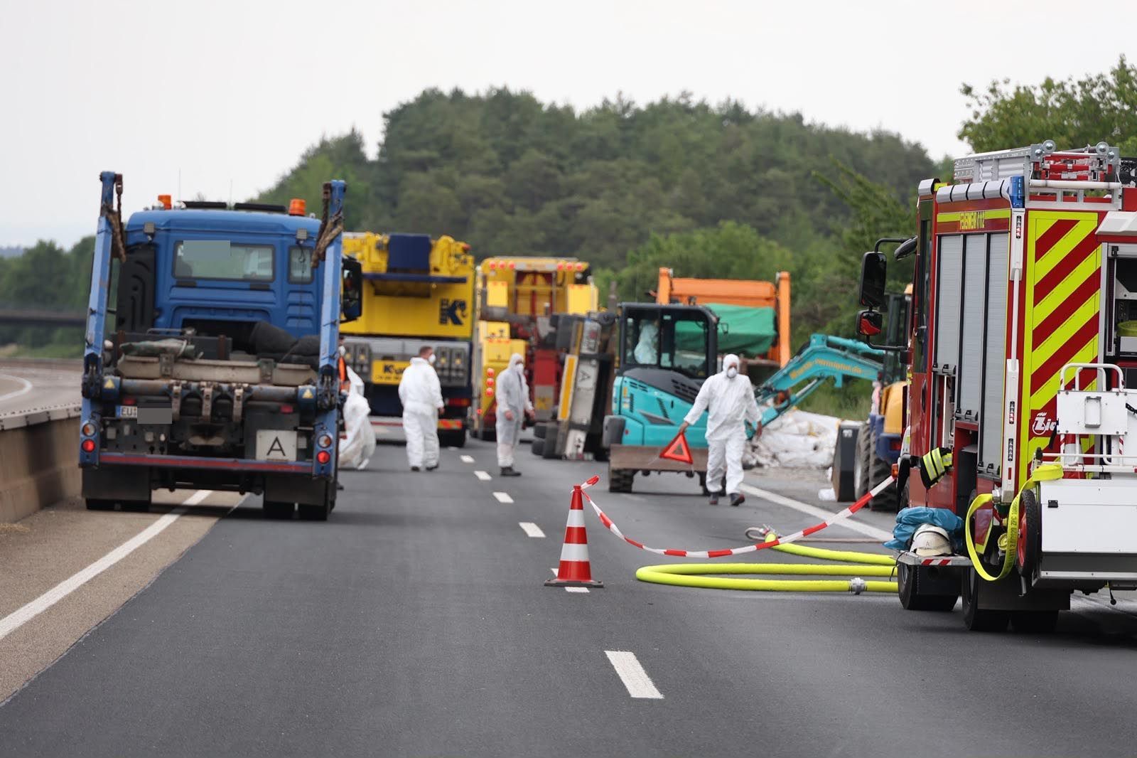 Einsatzkräfte in Schutzanzügen laufen über die Autobahn. Im Hintergrund ist der umgestürzte Anhänger zu sehen.
