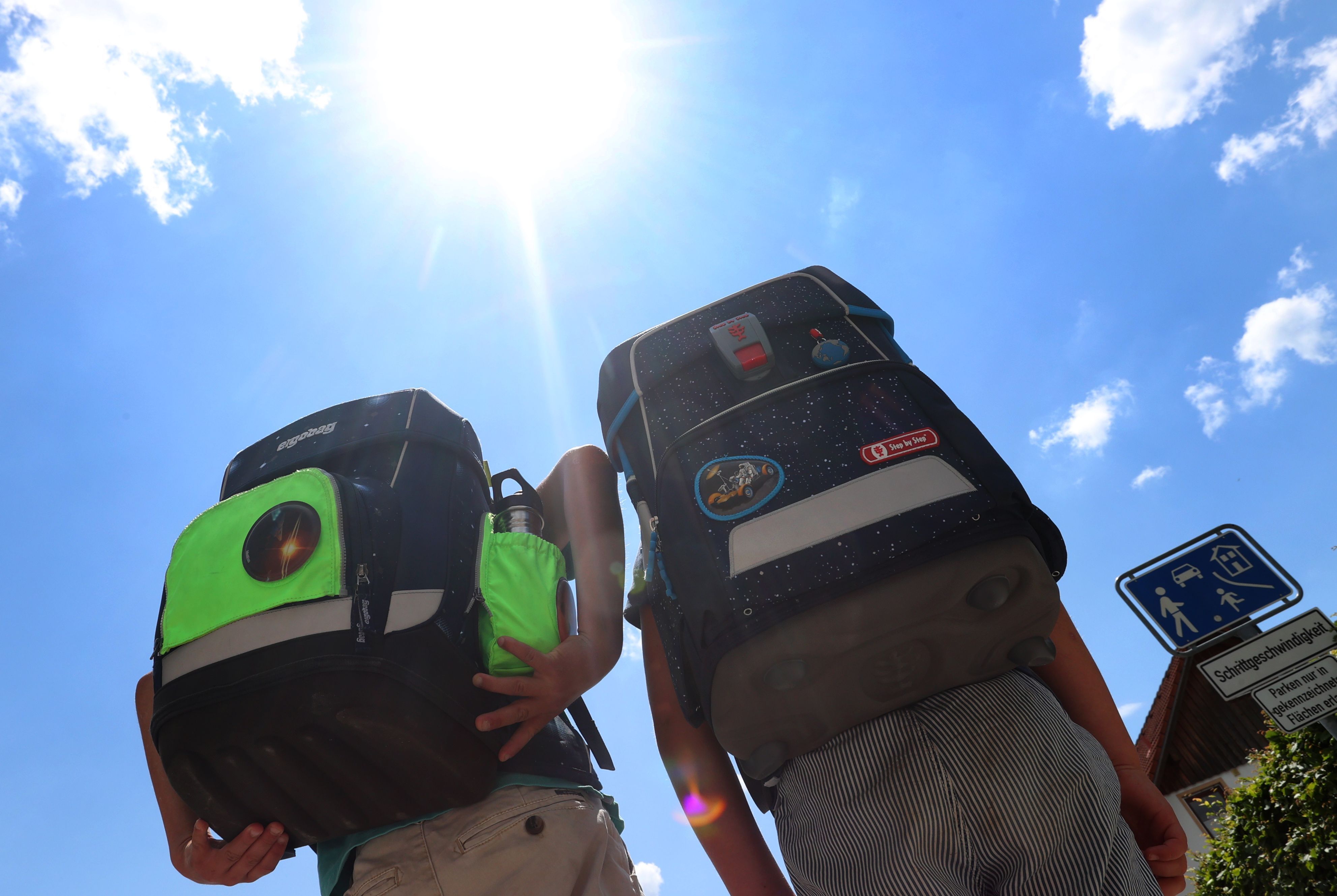 Zwei Kinder haben Hitzefrei und laufen mit ihren Büchertaschen im Sonnenschein nach Hause.
