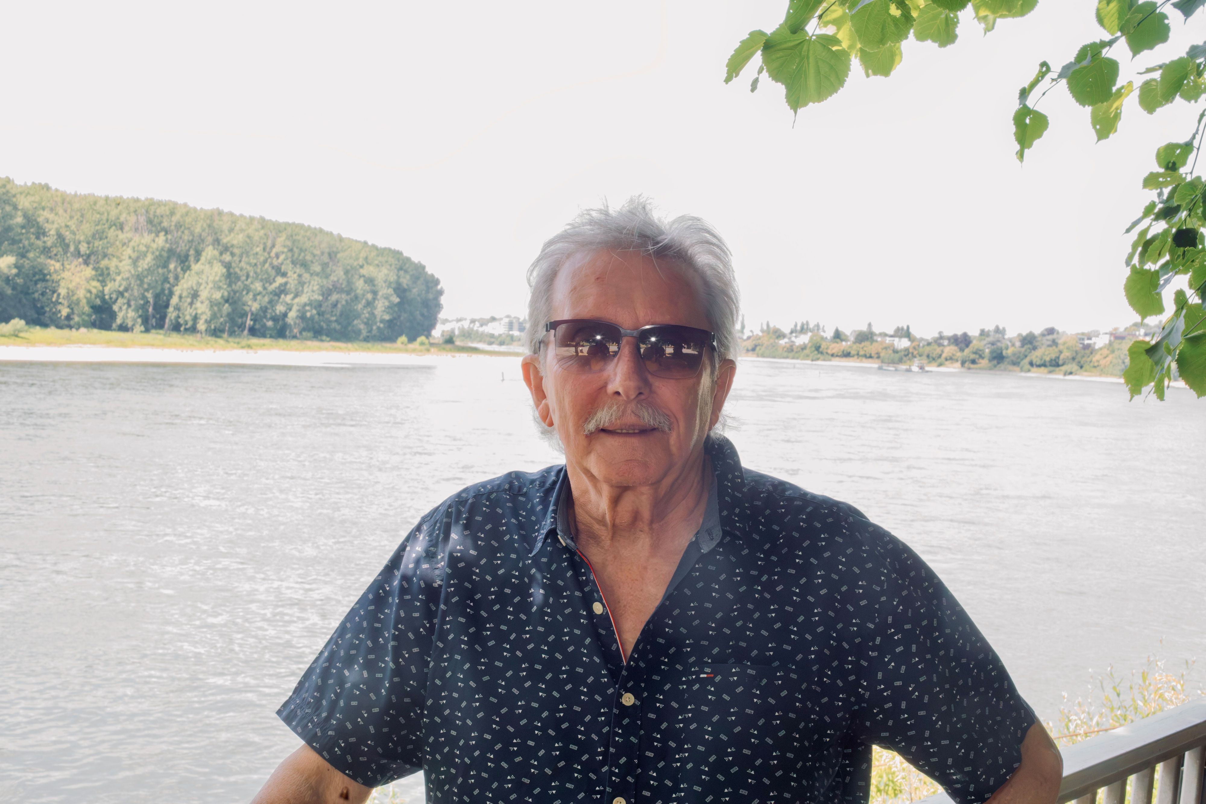 Ein Mann mit kurzem grauen Haar und Sonnenbrille und Schnurrbart steht vor dem Rhein in Porz.