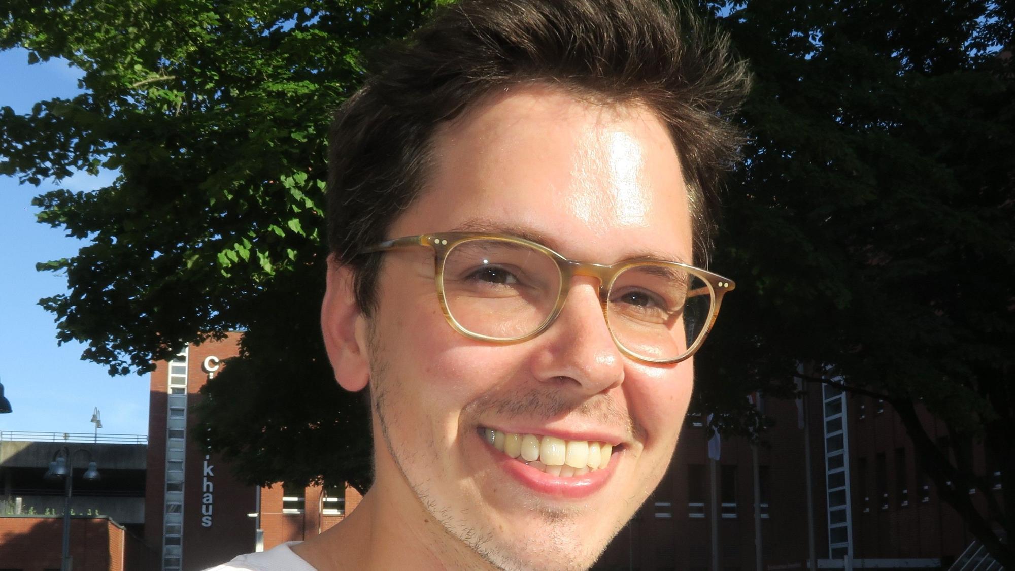 Ein junger Mann mit Brille und dunklem Haar blickt lachend in die Kamera.