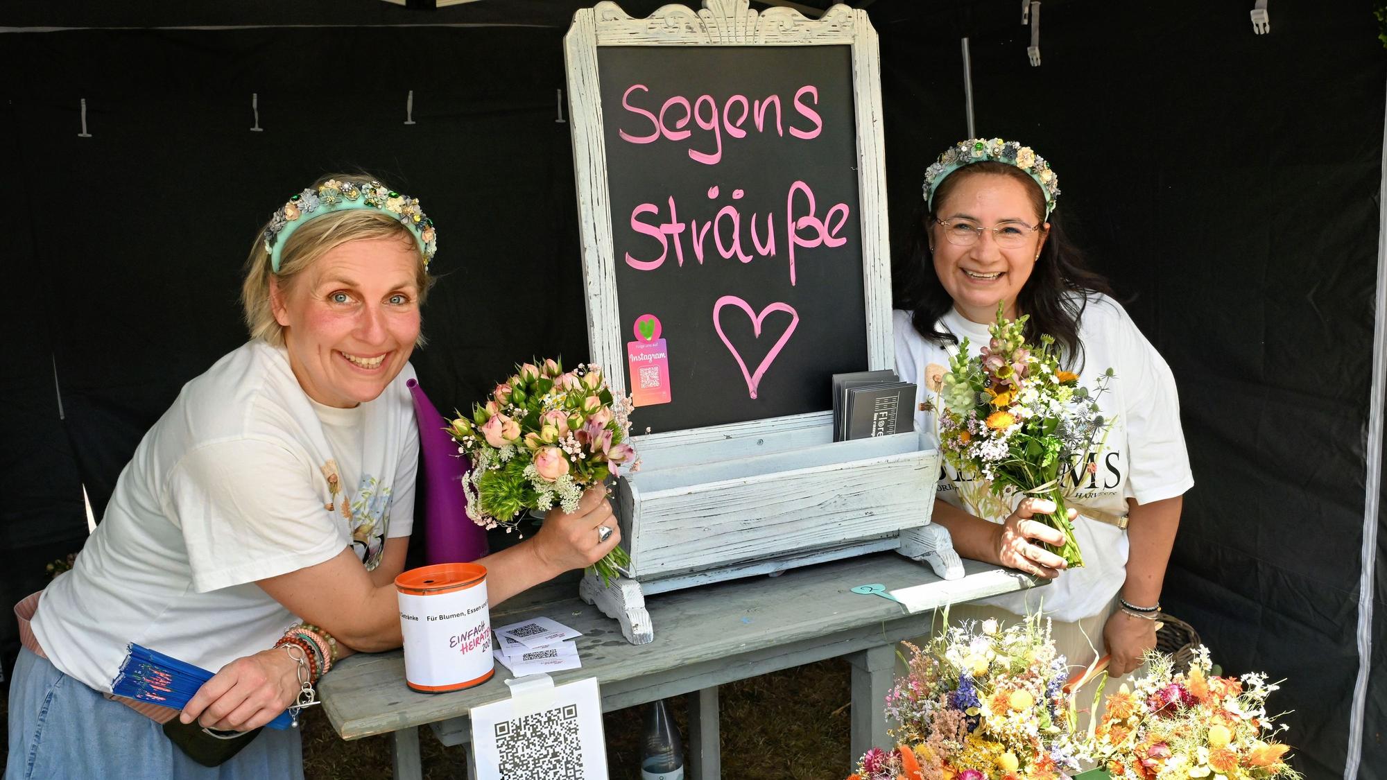 Die beiden Frauen mit Blumenkränzen im Haar halten Blumensträuße in die Kamera.