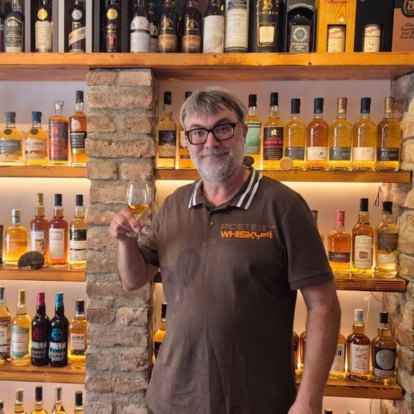 Ein Mann mit Bart und Brille steht vor einem Regal, gefüllt mit Whiskyflaschen, und hält ein Glas Whisky in der Hand.