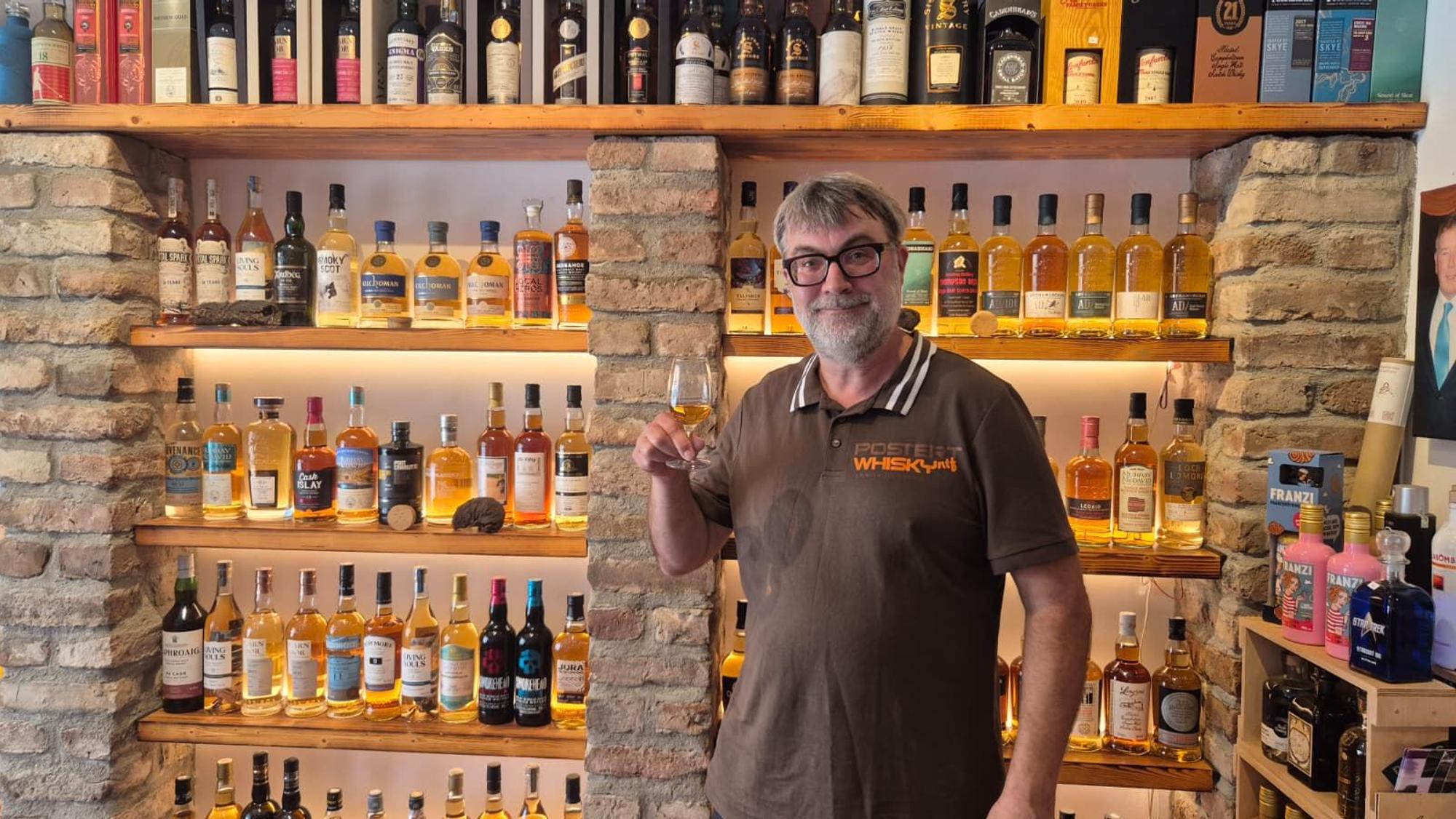 Ein Mann mit Bart und Brille steht vor einem Regal, gefüllt mit Whiskyflaschen, und hält ein Glas Whisky in der Hand.