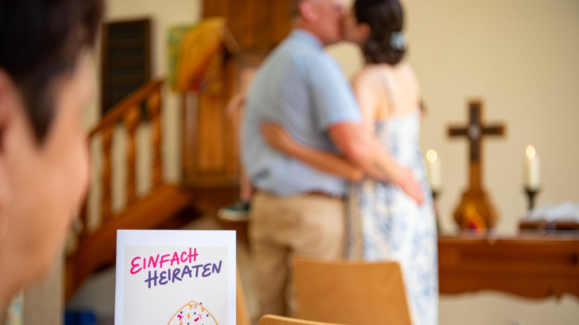 Eine Frau hält in Liederheft mit dem Schriftzug „Einfach Heiraten“ in der Hand, im Hintergrund ist ein Brautpaar zu sehen, das sich küsst.