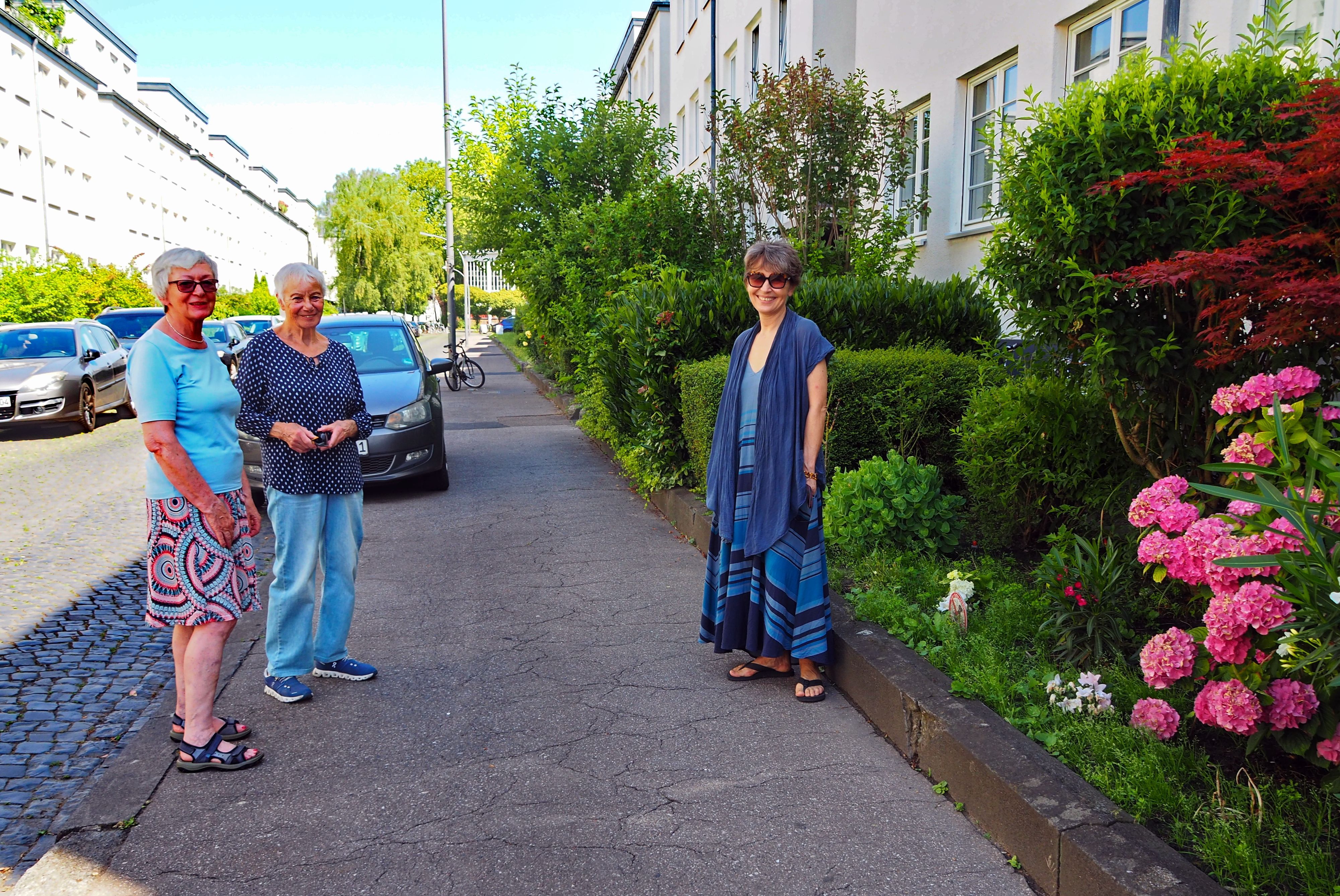 Drei Frauen stehen auf einem Bürgersteig. Ein Auto parkt hinter ihnen.