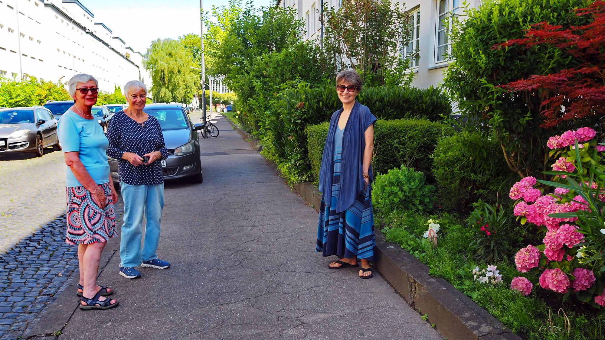 Drei Frauen stehen auf einem Bürgersteig. Ein Auto parkt hinter ihnen.
