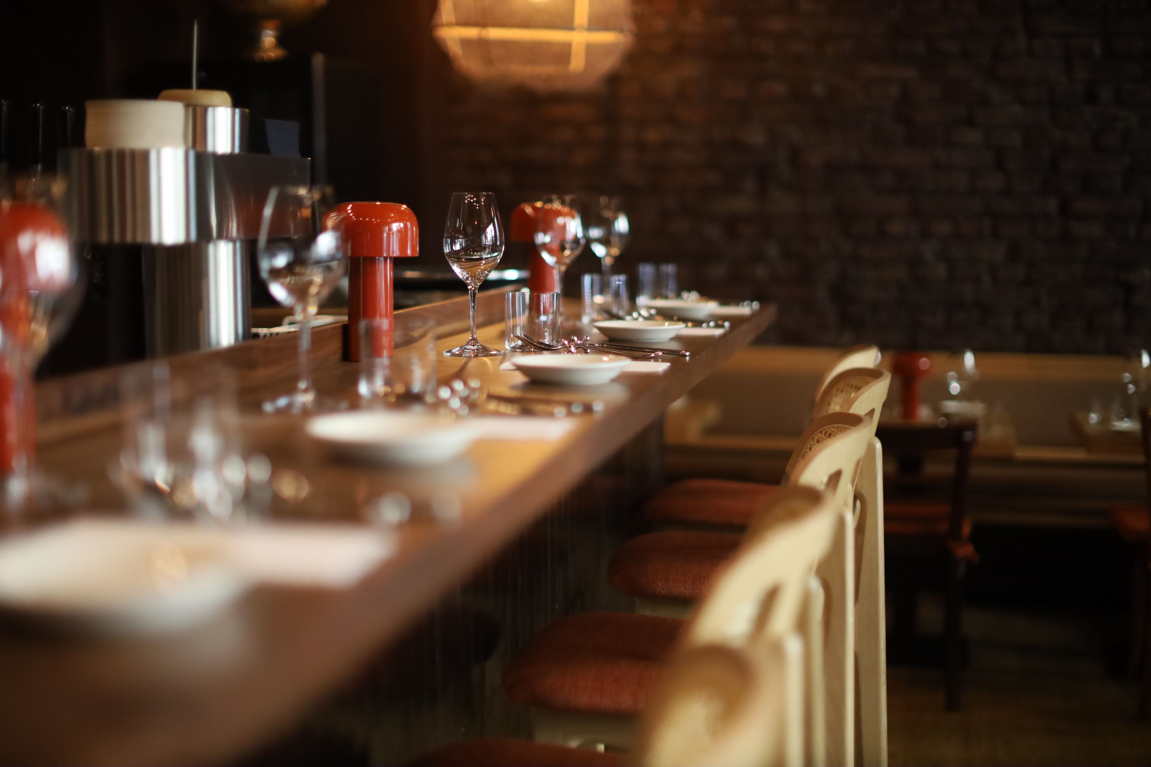 Die eingedeckte Bar: Auf dem Tisch stehen Gläser, Teller und Tischlampen, zudem helle Barstühle