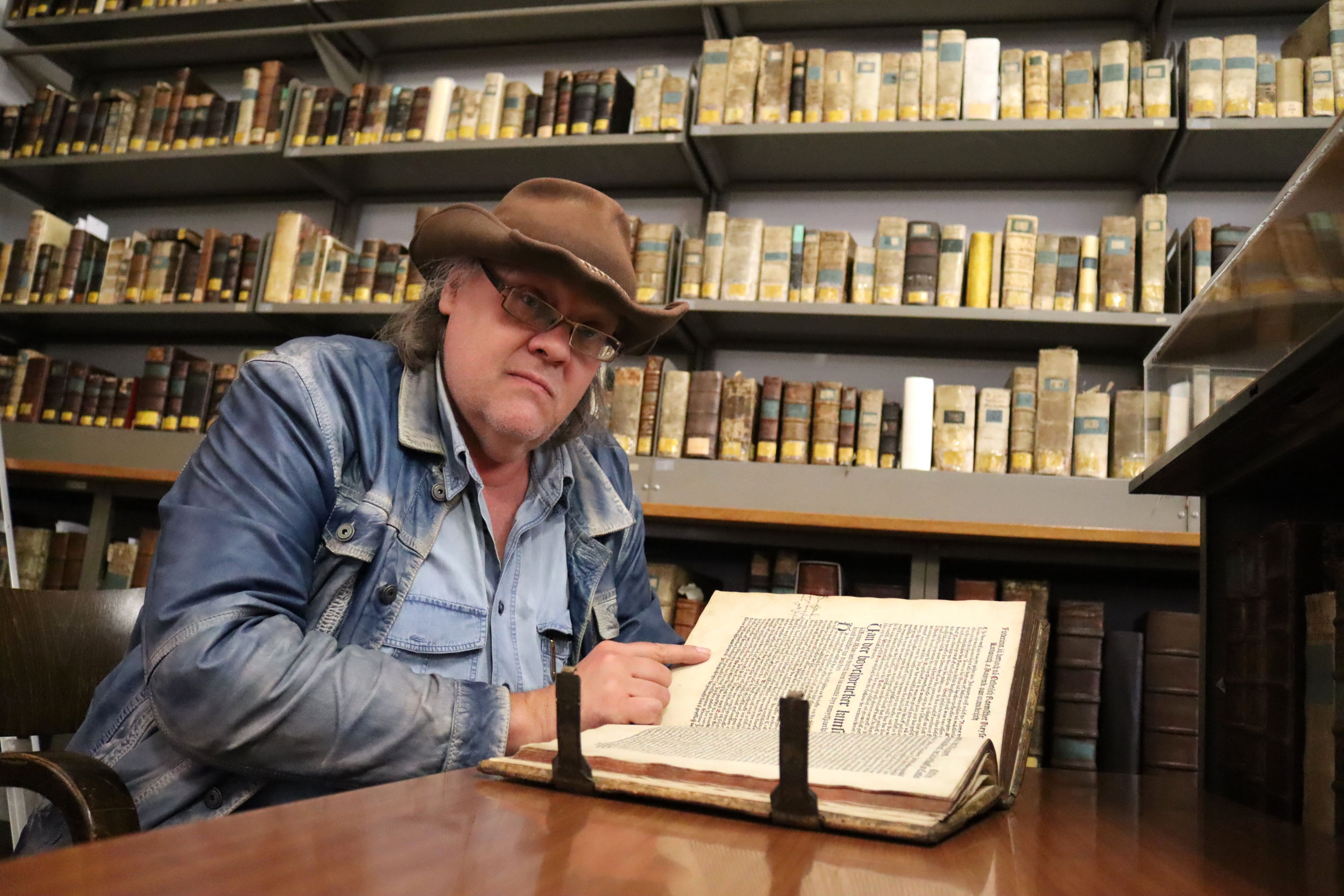 Bongart, der einen Hut trägt, sitzt an einem Tisch, vor sich ein Buch. Im Hintergrnd Regale.