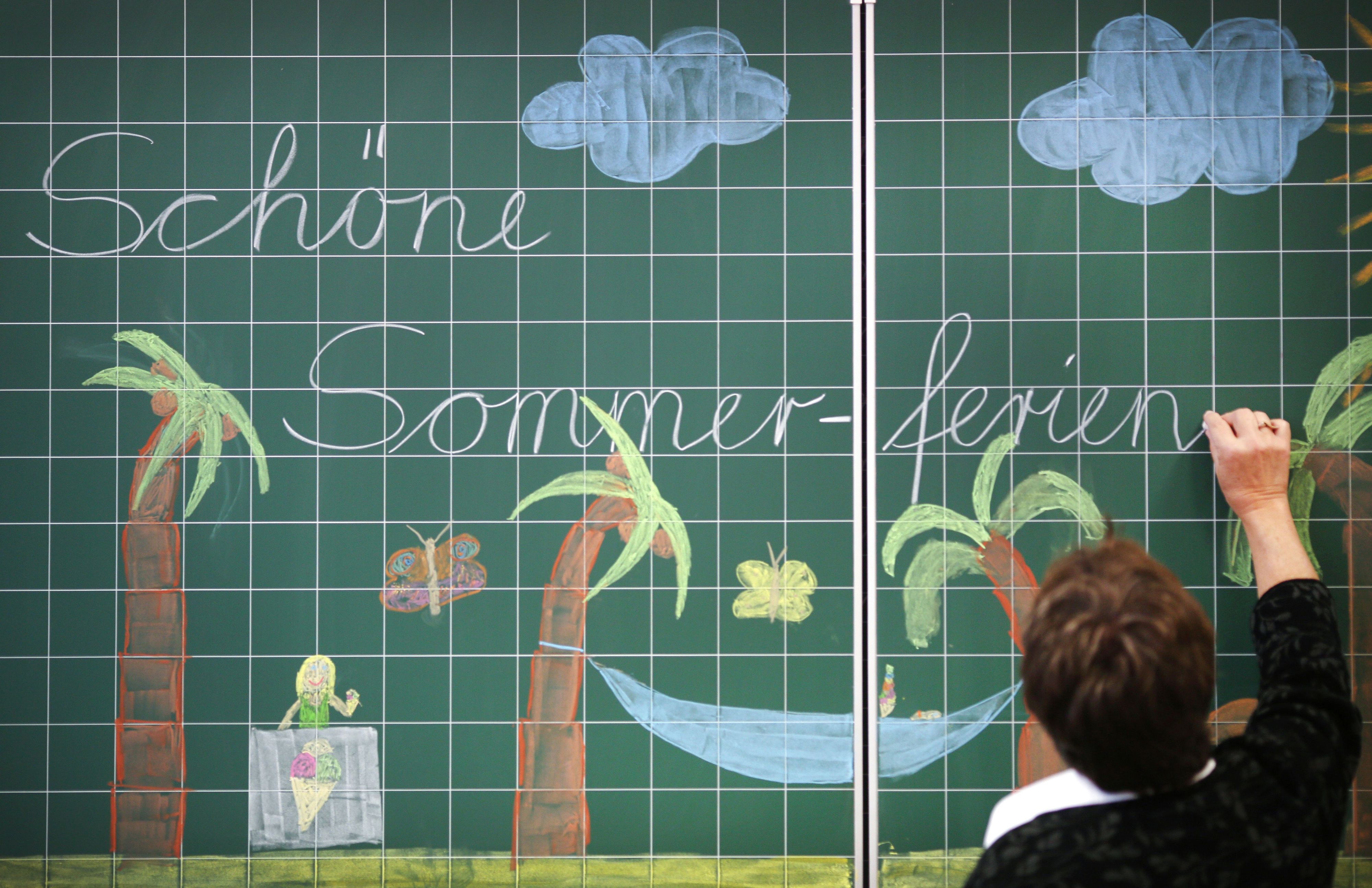 Eine Lehrerin schreibt „Schöne Sommer-ferien“ an eine mit Palmen bemalte Tafel in einer Grundschule.