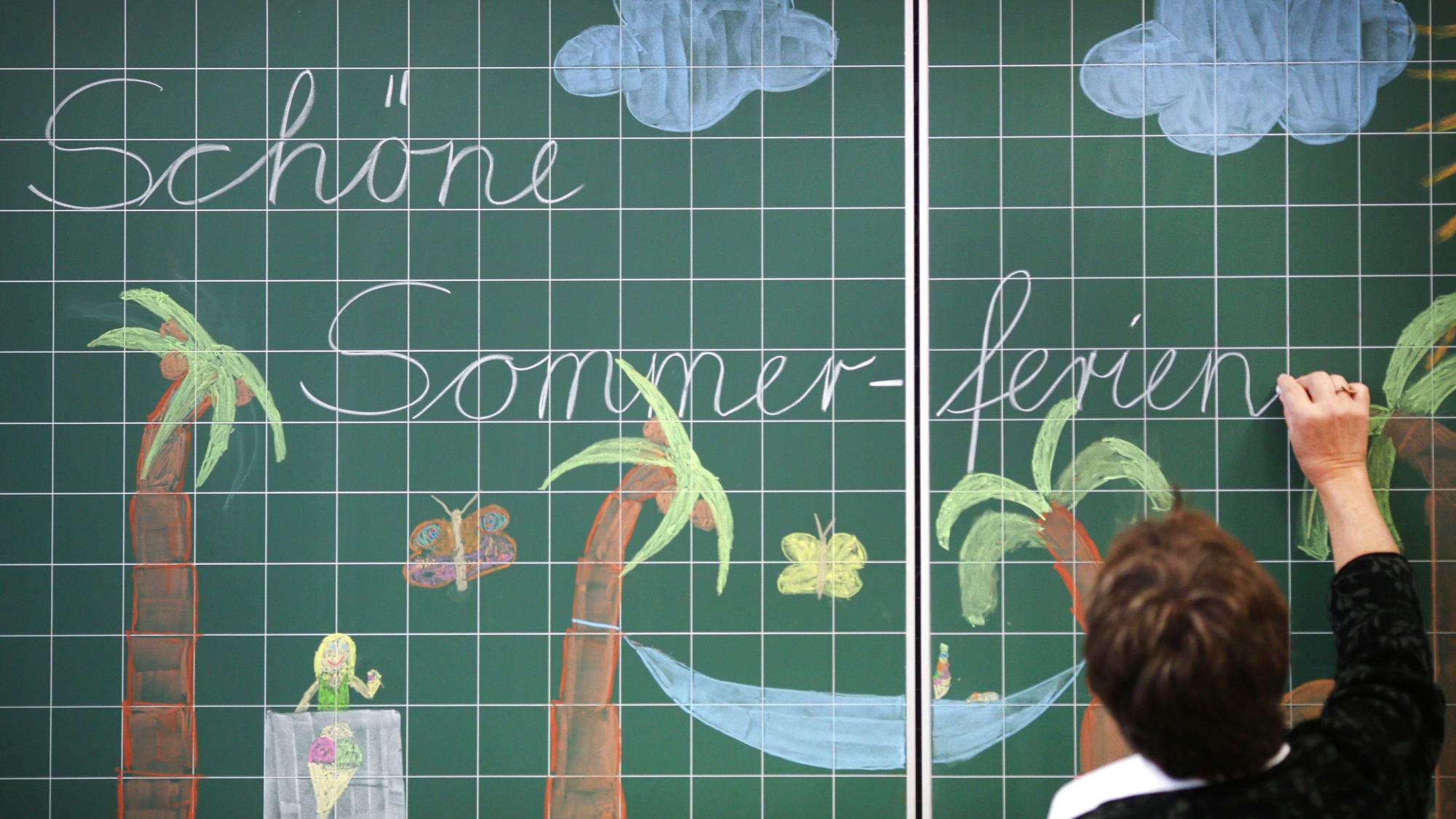 Eine Lehrerin schreibt „Schöne Sommer-ferien“ an eine mit Palmen bemalte Tafel in einer Grundschule.
