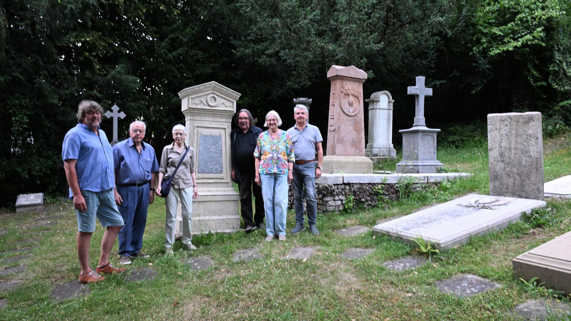 Vier Männer und zwei Frauen stehen auf einem Friedhof vor uralten Grabsteinen udn Grabplatten. Dazwischen Wiese.