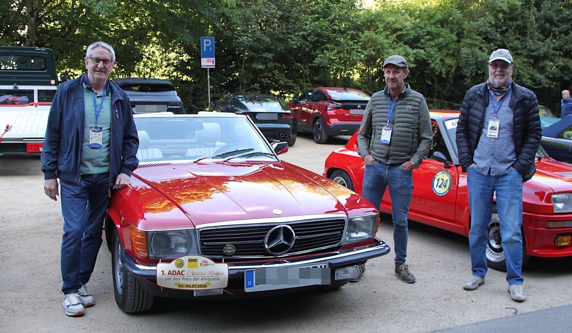 Die drei Männer stehen neben einem roten Mercedes.