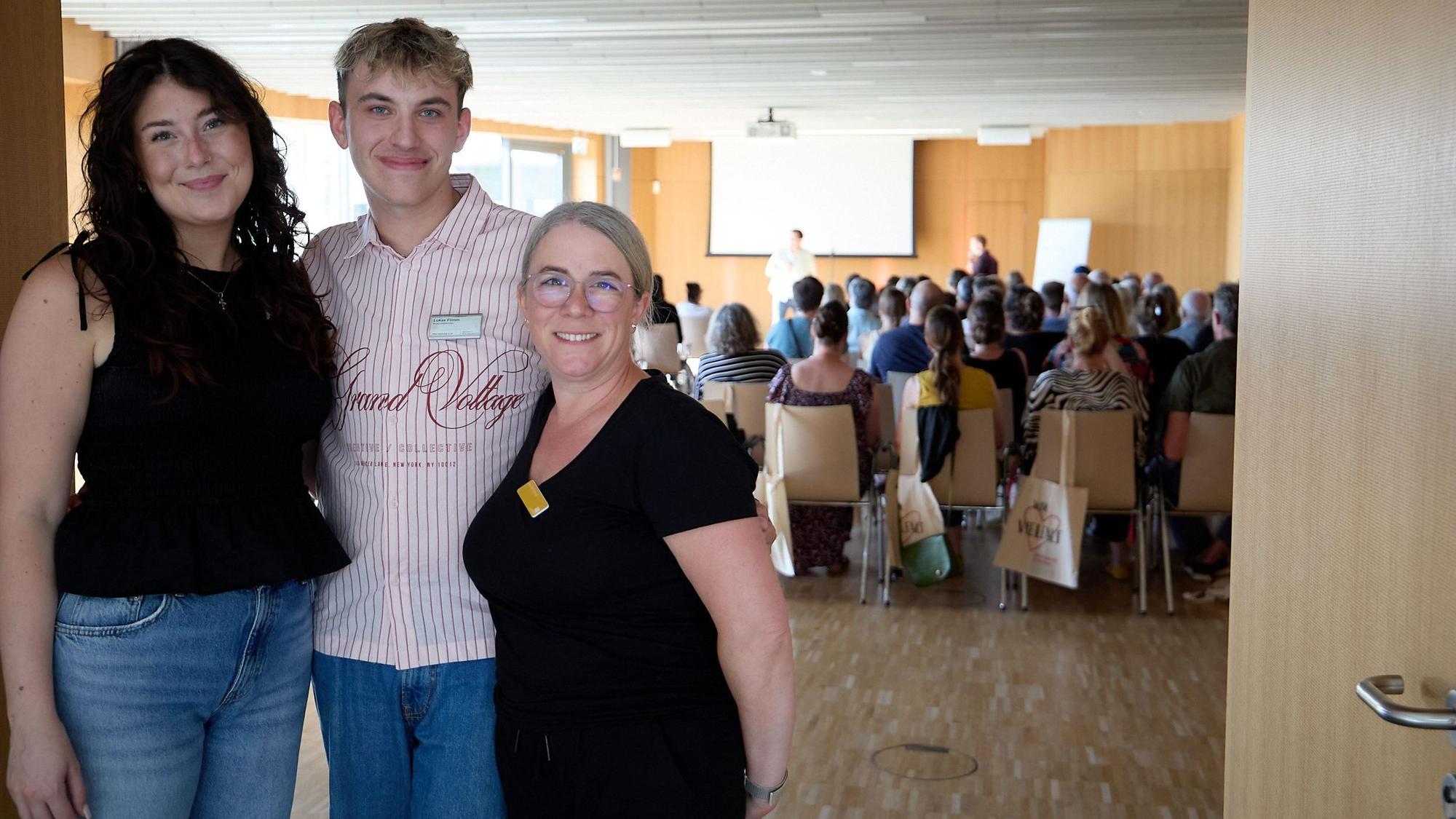 Lukas Flimm, Jennifer Wenck (rechts) und Anna-Lena Stockem stehen vor einem Saal, in dem viele Leute sitzen.