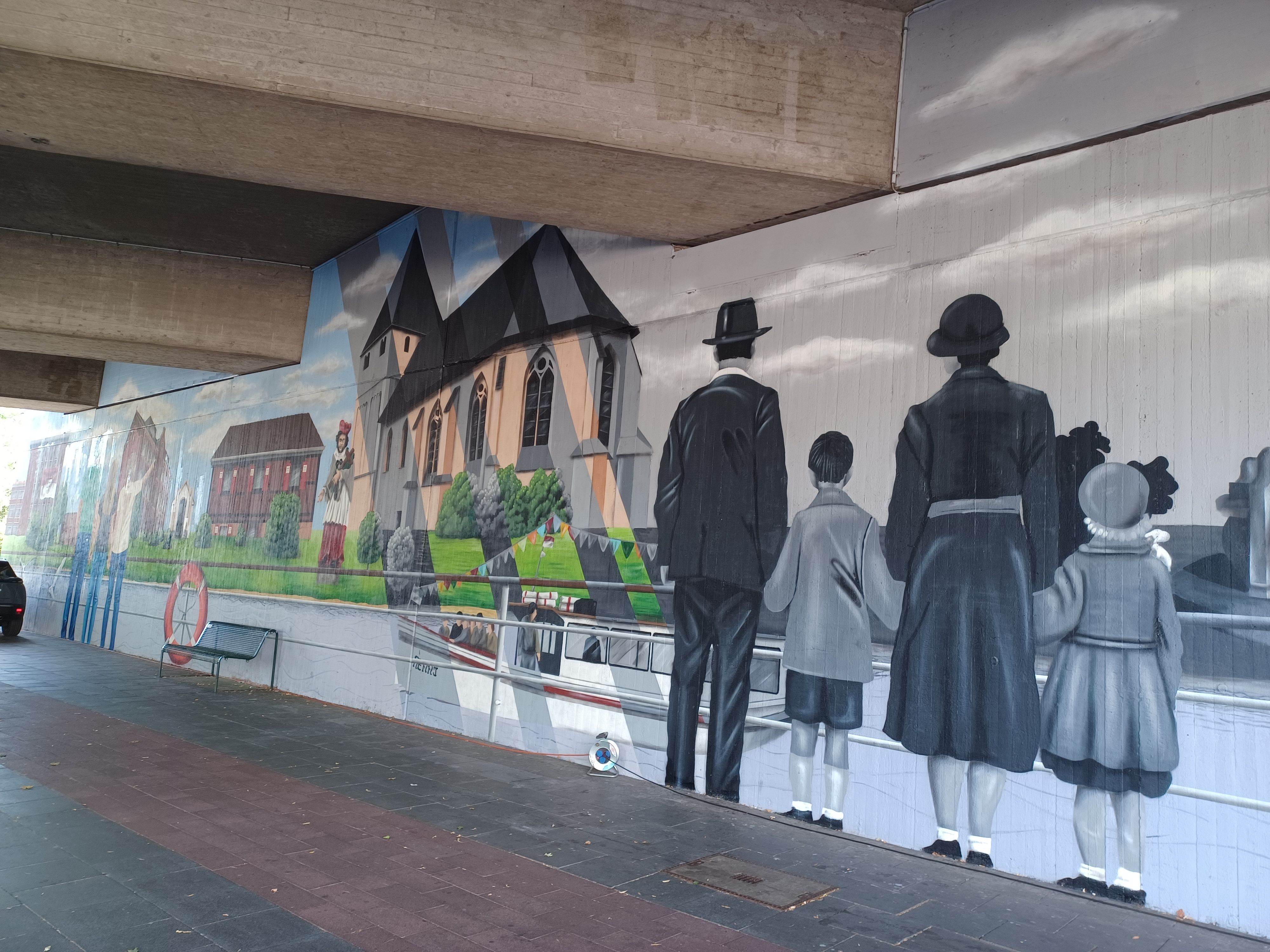 Die eine Seite einer Tunnelunterführung ist mit einem Rheinpanorama verziert. Gebäude aus Niehl sind zu sehen, Schiffe und eine Familie, die auf den Rhein blickt.