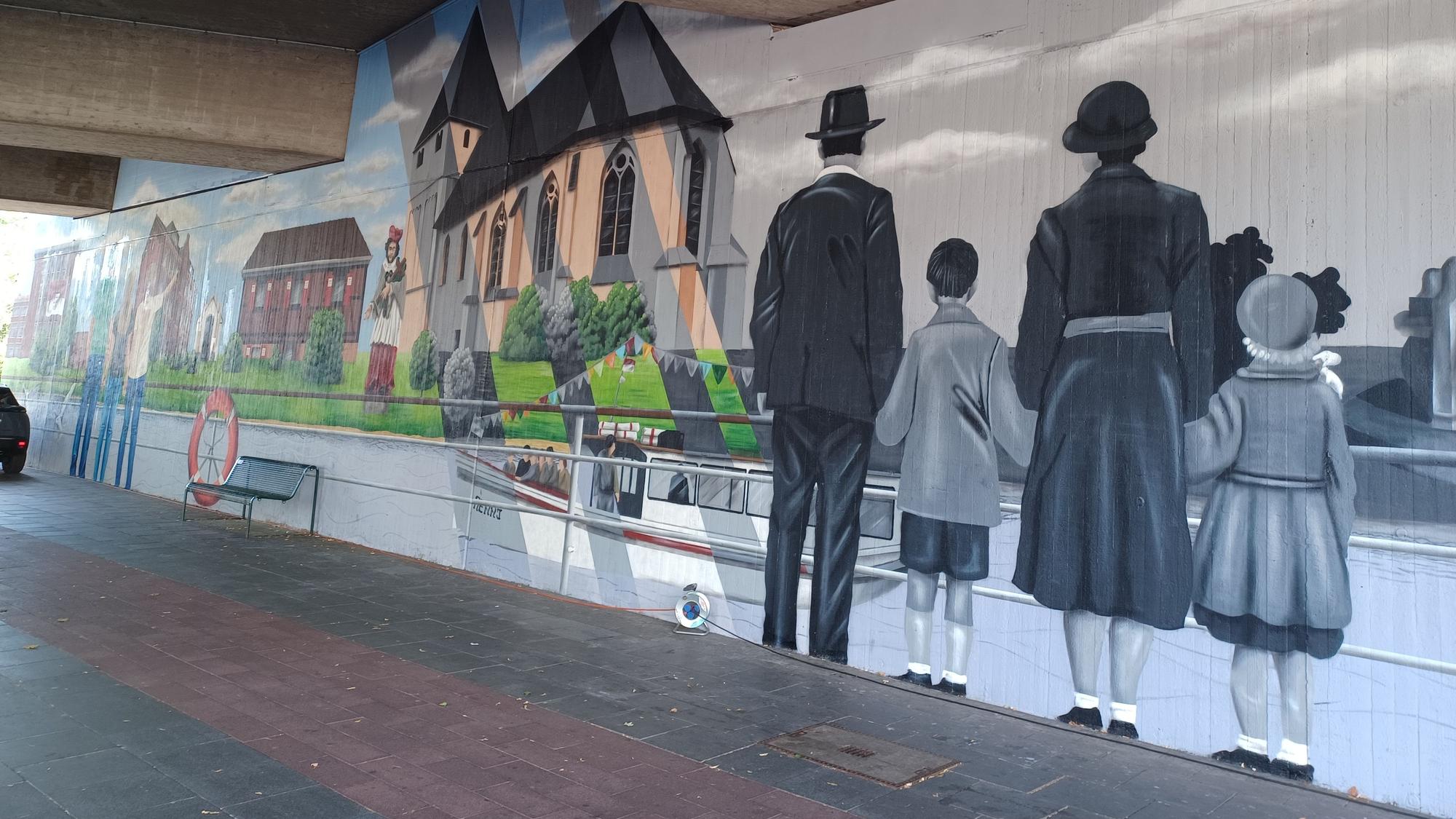 Die eine Seite einer Tunnelunterführung ist mit einem Rheinpanorama verziert. Gebäude aus Niehl sind zu sehen, Schiffe und eine Familie, die auf den Rhein blickt.