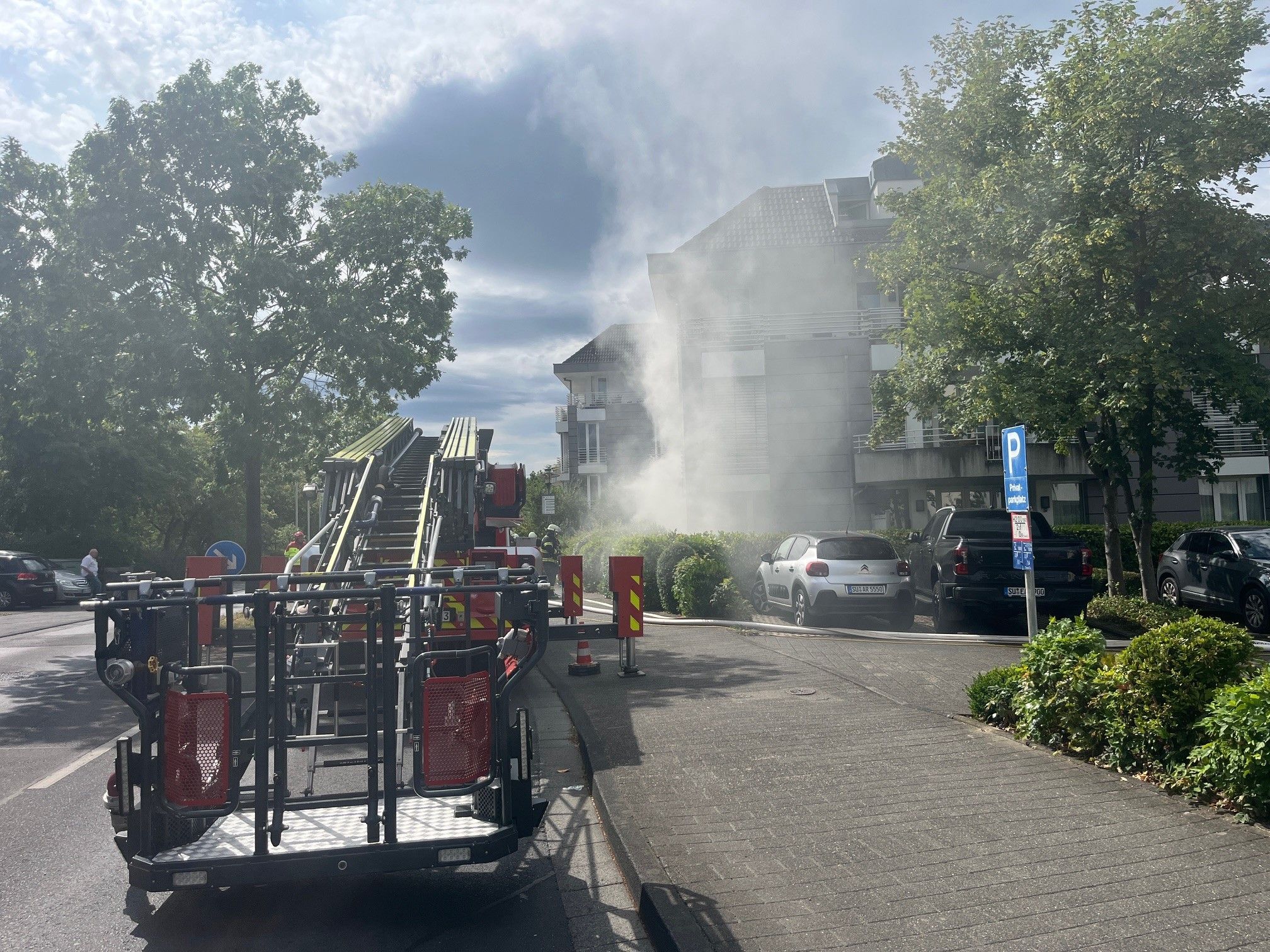 Ein Drehleiterfahrzeug der Feuerwehr. Im Hintergrund dringt Rauch aus dem Keller eines Gebäudes.
