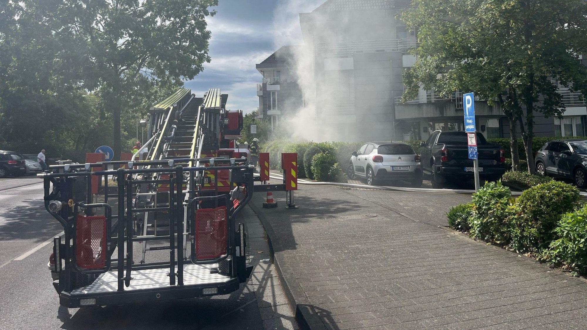 Ein Drehleiterfahrzeug der Feuerwehr. Im Hintergrund dringt Rauch aus dem Keller eines Gebäudes.