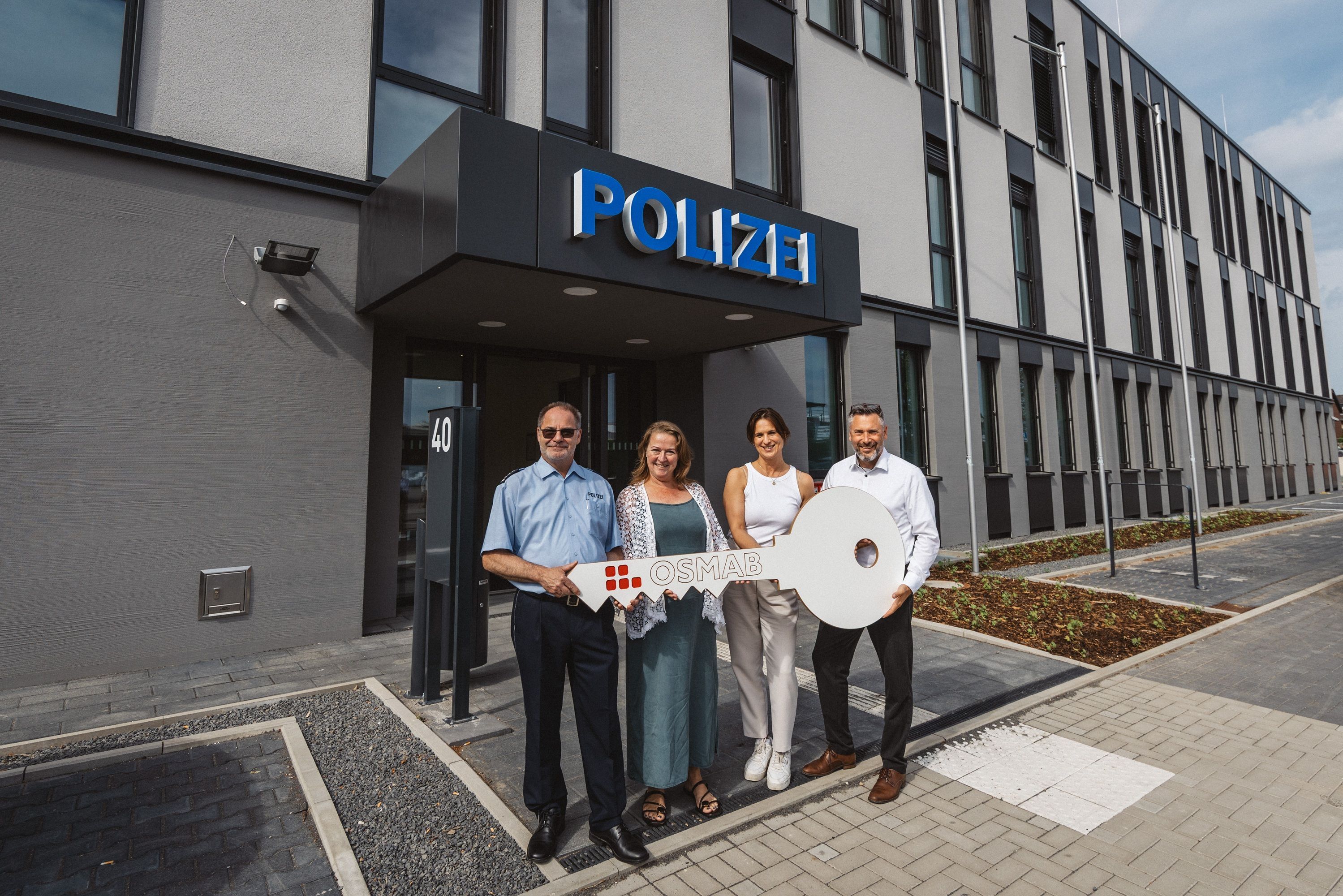 Auf dem Bild sind zwei Frauen und zwei Männer vor dem neuen Polizeigebäude zu sehen, sie halten einen großen symbolischen Schlüssel in der Hand.