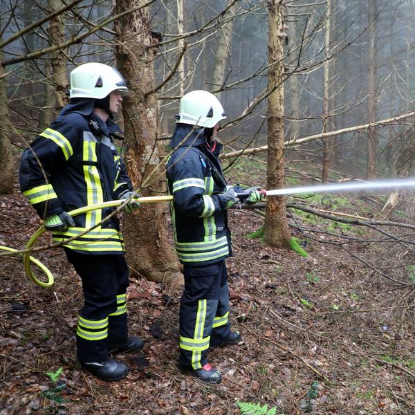 Zu sehen sind zwei Feuerwehrmänner mit einem Schlauch in einem Wald.