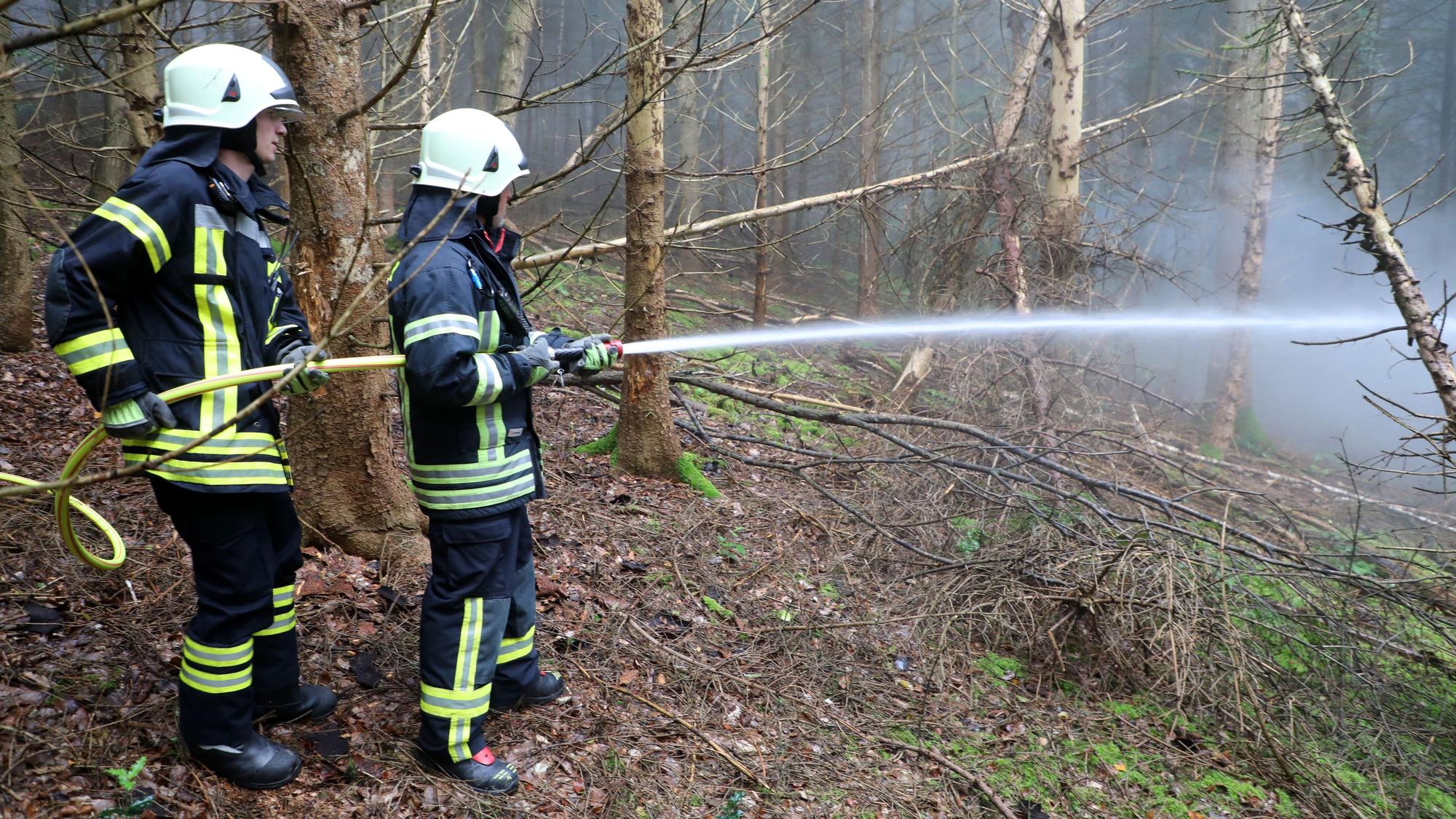 Zu sehen sind zwei Feuerwehrmänner mit einem Schlauch in einem Wald.