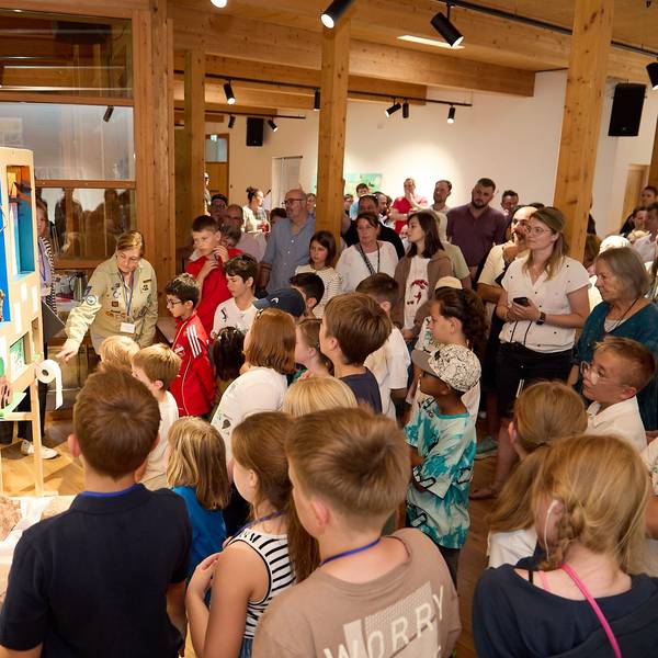 Viele Kinder und einige Erwachsene drängen sich um eine Schautafel. Links steht eine Frau, die in ein Mikrofon spricht.