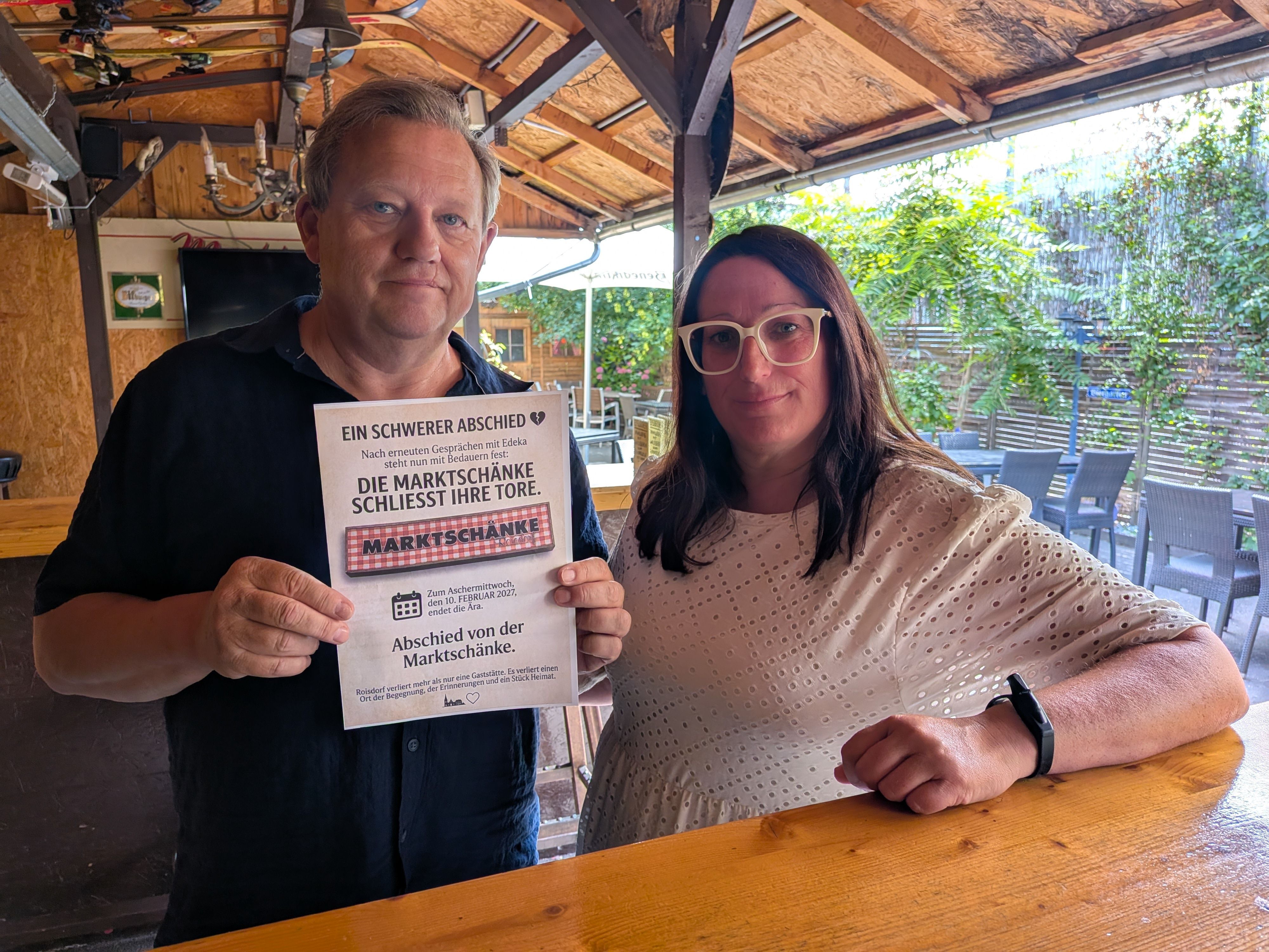 Stephan Kempf und Andrea May stehen im Lokal. Kempf hält einen Zettel vor sich, auf dem „Die Marktschänke schließt ihre Tore“ steht.