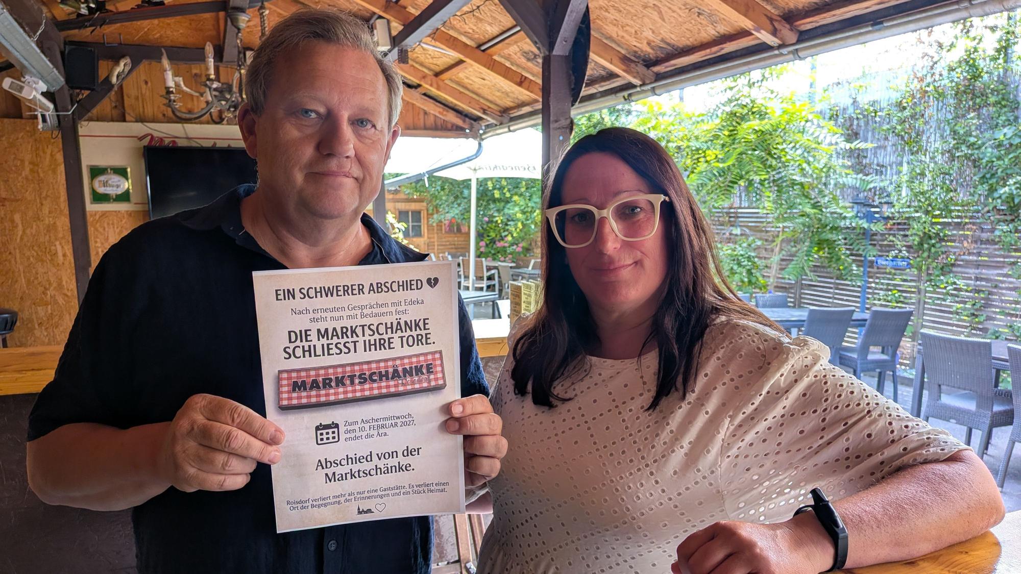 Stephan Kempf und Andrea May stehen im Lokal. Kempf hält einen Zettel vor sich, auf dem „Die Marktschänke schließt ihre Tore“ steht.