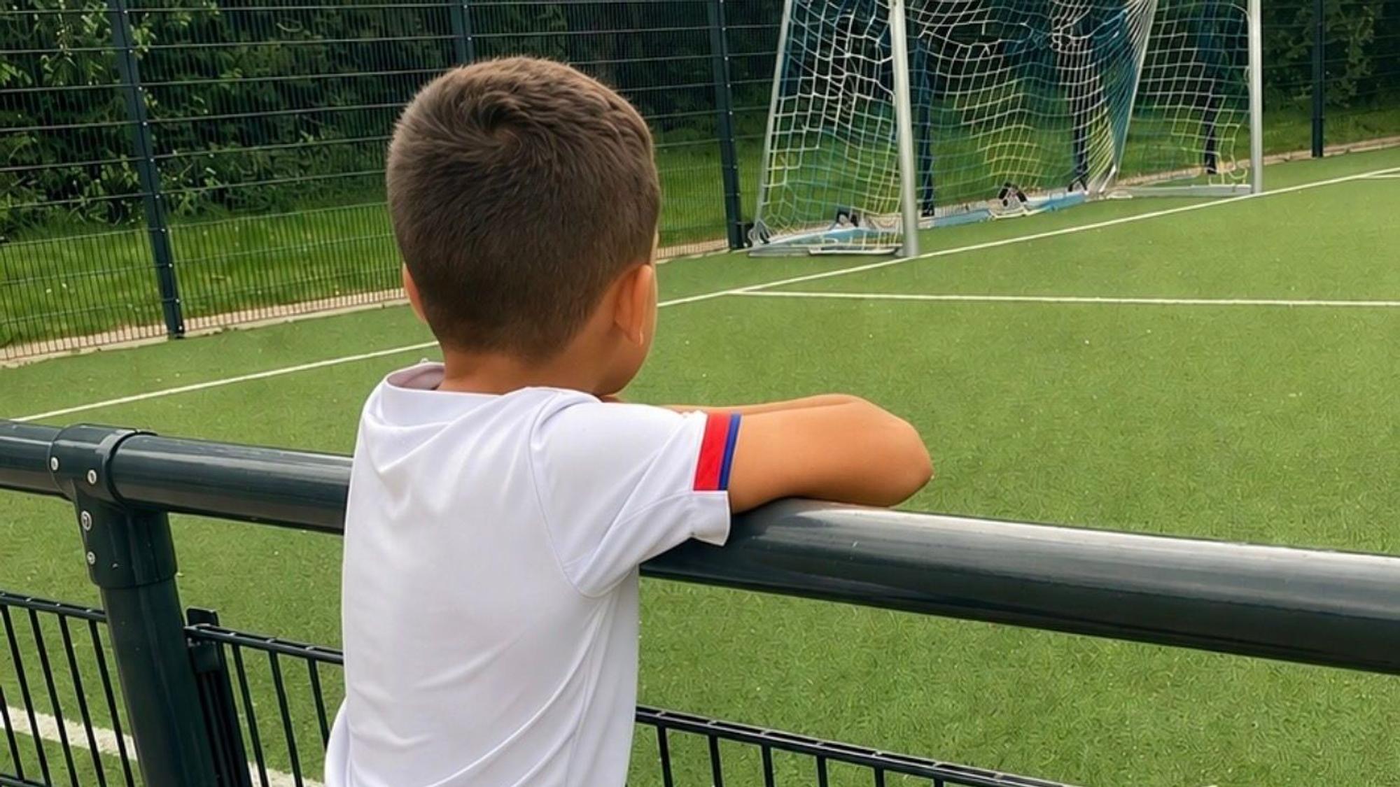 Junge schaut über einen Zaun auf einen Fußballplatz.