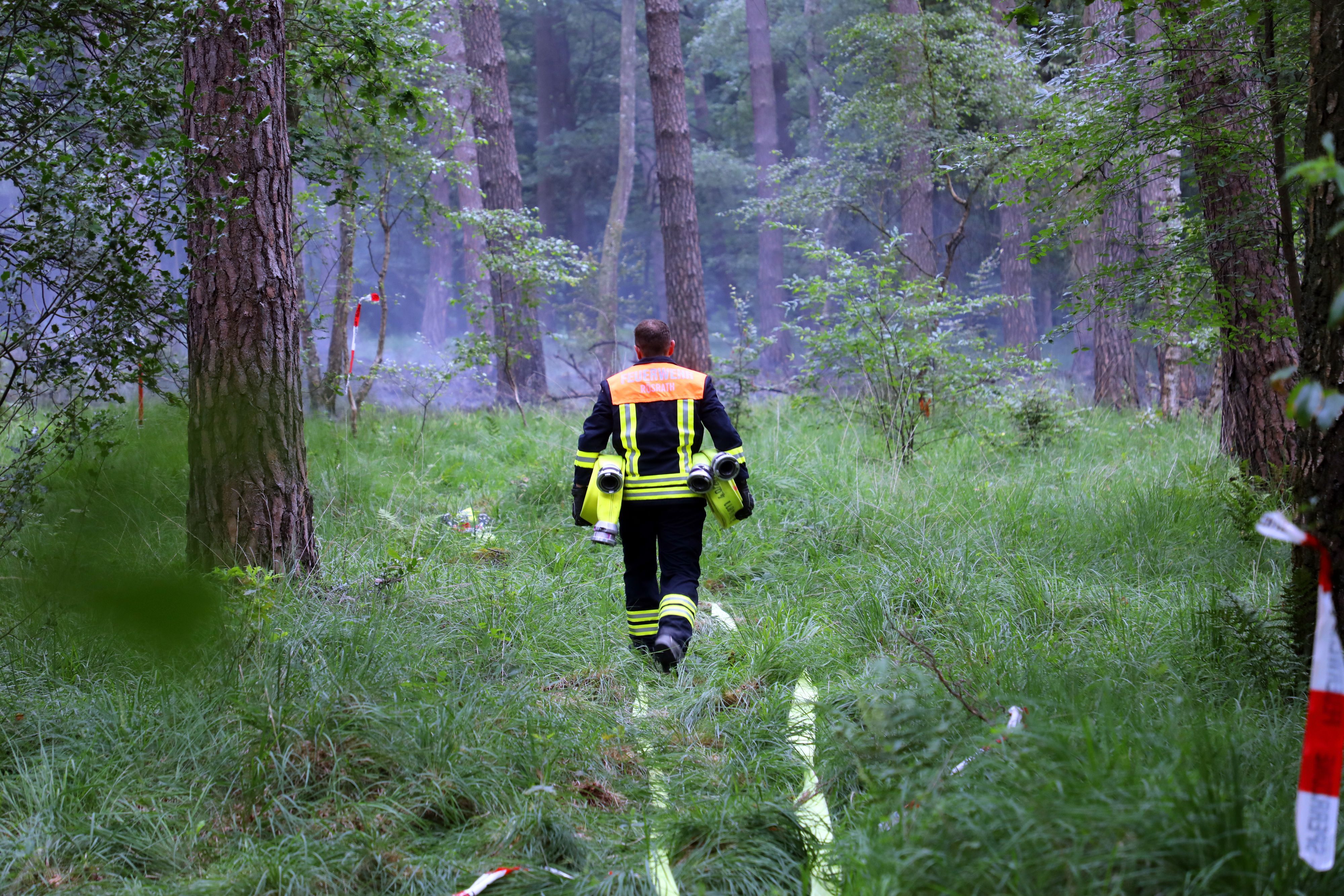 Ein Feuerwehrmann trägt Schläuche in einen  Wald. Im Hintergrund ist Qualm zu sehen.