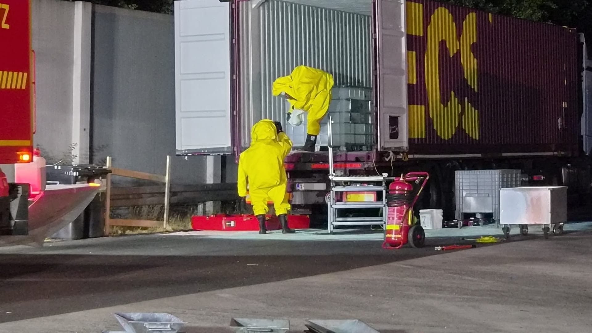 Das Bild zeigt zwei Feuerwehrleute, die unter großen Vorsichtsmaßnahmen die Container untersuchen.
