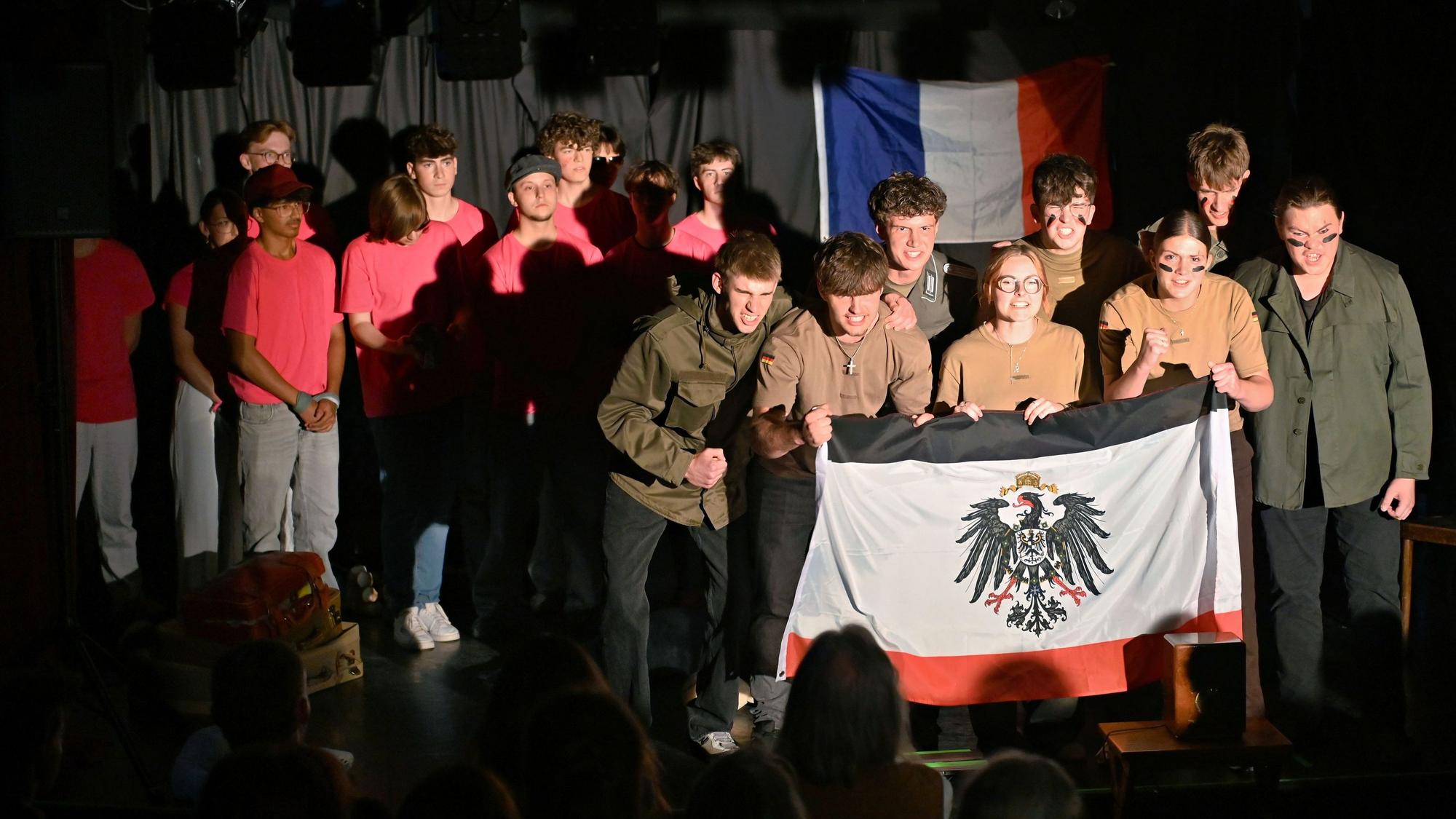 Die Schülerinnen und Schüler bilden auf der Bühne zwei Gruppen. Die einen tragen T-Shirts in Pink, die anderen sind in Oliv gekleidet und zeigen eine Flagge mit dem Reichsadler.