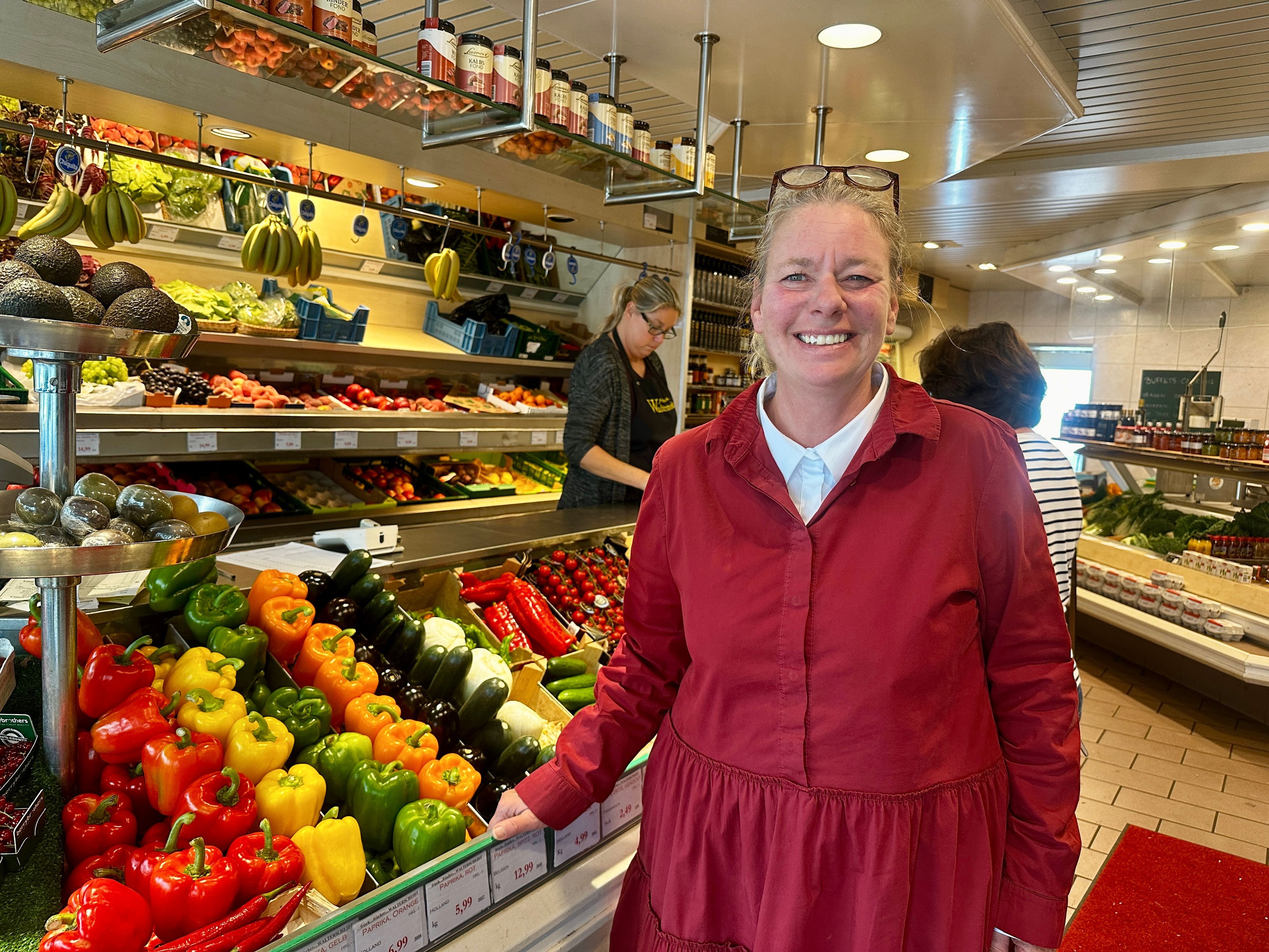 Eine Frau in roter Jacke mit der hochgeschobenen Brille im Haar steht neben einem Aufsteller mit Obst und Gemüse.