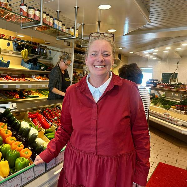 Eine Frau in roter Jacke mit der hochgeschobenen Brille im Haar steht neben einem Aufsteller mit Obst und Gemüse.