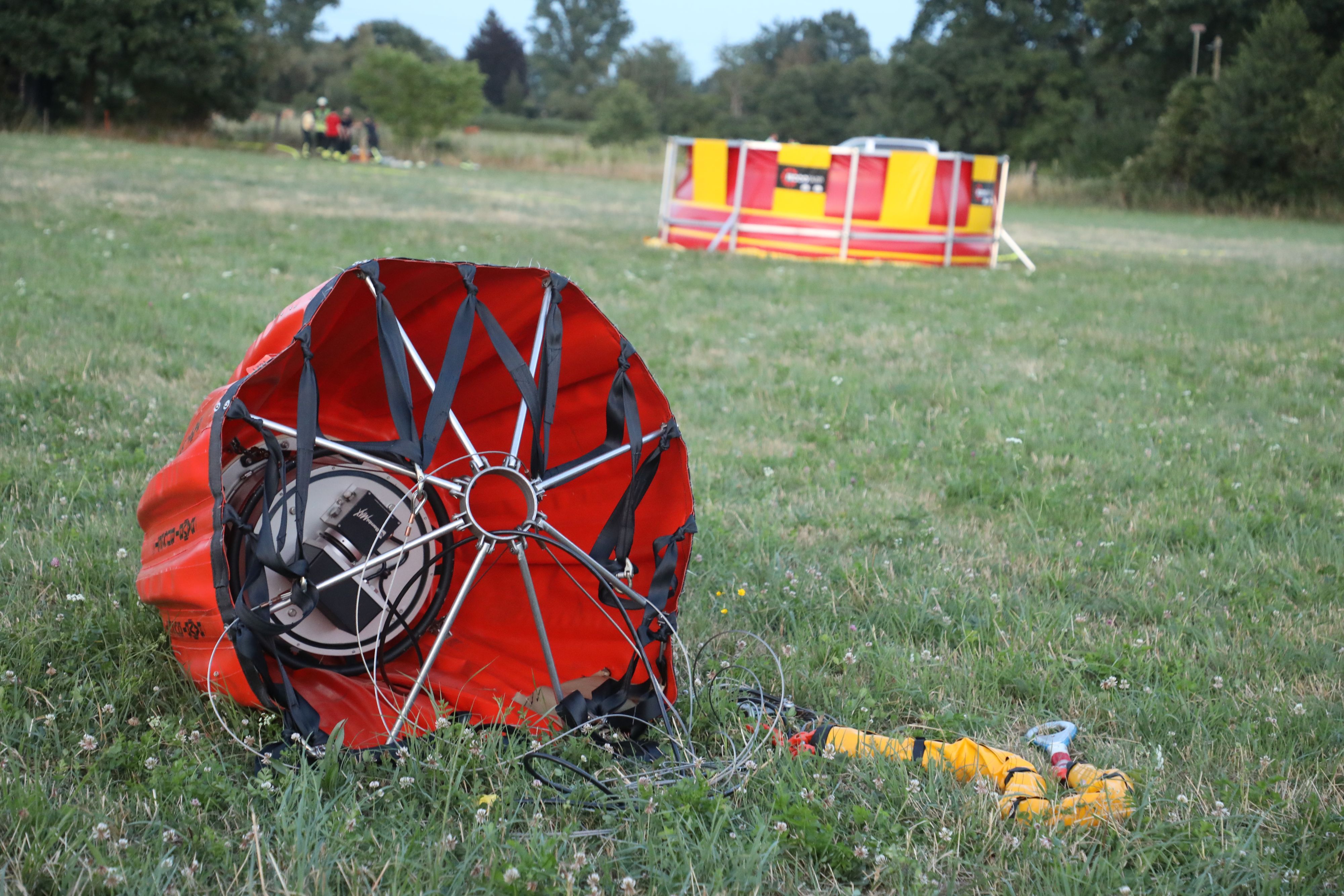 Ein „Bambi Bucket“ liegt im Vordergrund auf einer Wiese. Ein Polizeihubschrauber bringt es mit Wasser gefüllt über das brennende Waldgebiet. Im Hintergrund ist das Faltbecken zu sehen, aus dem nachgefüllt wird.