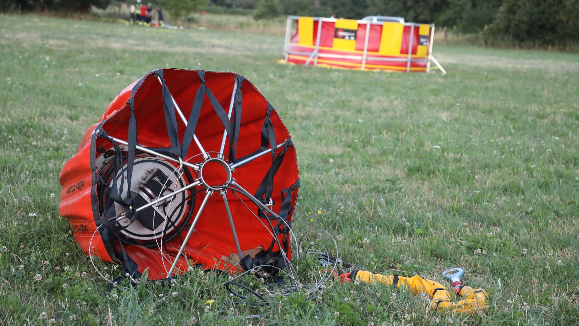 Ein „Bambi Bucket“ liegt im Vordergrund auf einer Wiese. Ein Polizeihubschrauber bringt es mit Wasser gefüllt über das brennende Waldgebiet. Im Hintergrund ist das Faltbecken zu sehen, aus dem nachgefüllt wird.