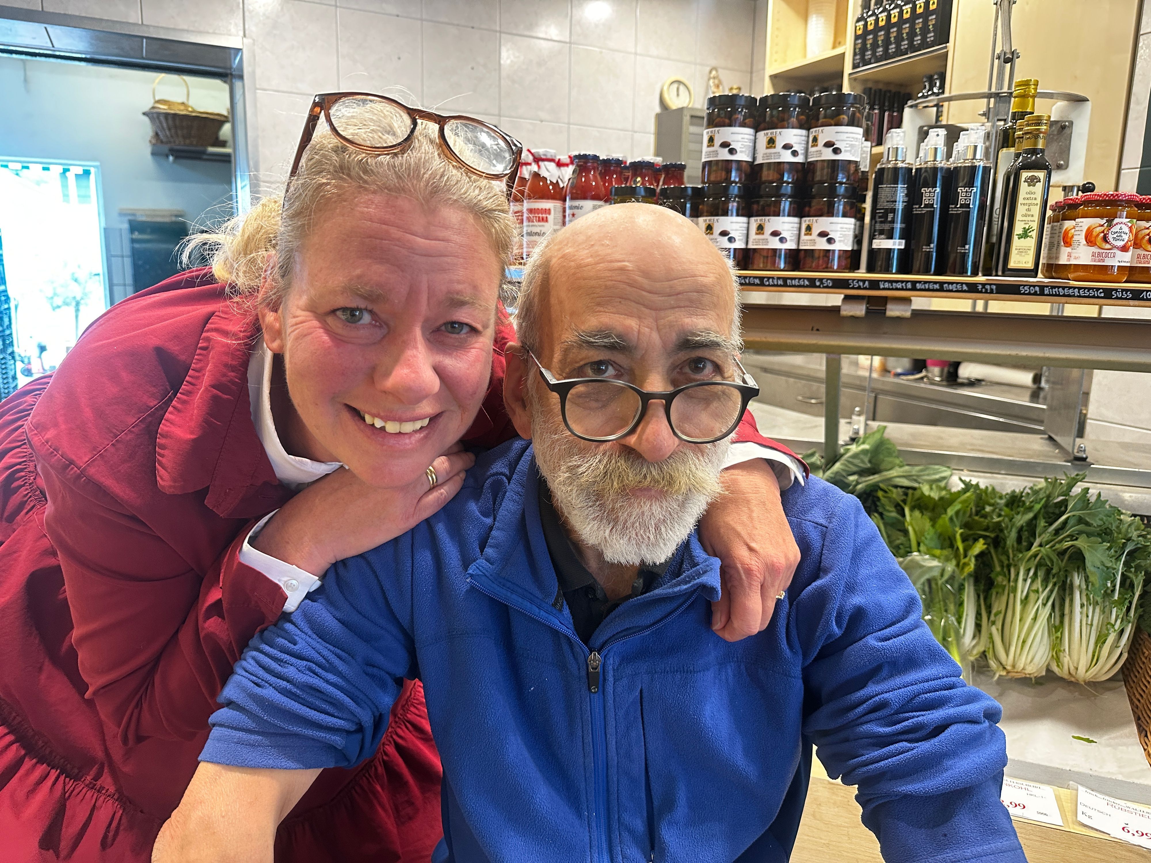 Eine Frau mit roter Jacke und Brille im Haar umarmt einen älteren Mann mit Bart, blauer Jacke und Brille.