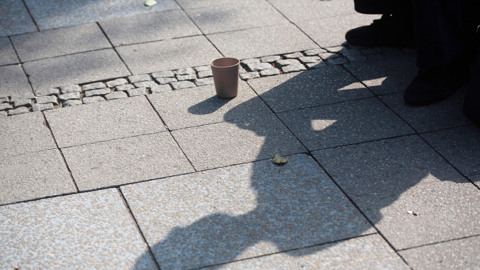 SYMBOLBILD - 10.07.2023, Berlin: Der Schatten einer Person mit einem Becher für Spenden fällt auf den Gehweg. (zu dpa: «Wohnungslosenhilfe fordert Trinkwasserstellen für Obdachlose») Foto: Fernando Gutierrez-Juarez/dpa +++ dpa-Bildfunk +++