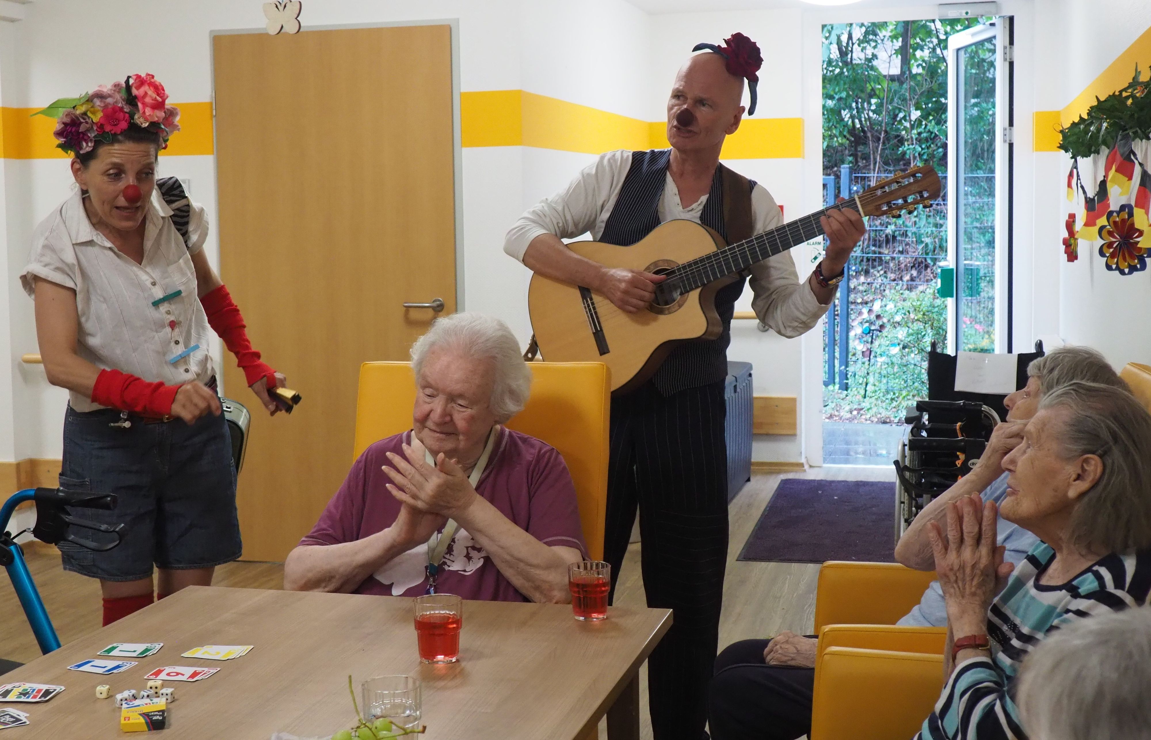 Ein Clown spielt Gitarre, ein weiblicher Clown links im Bild singt für die Seniorinnen, die begeistert klatschen.