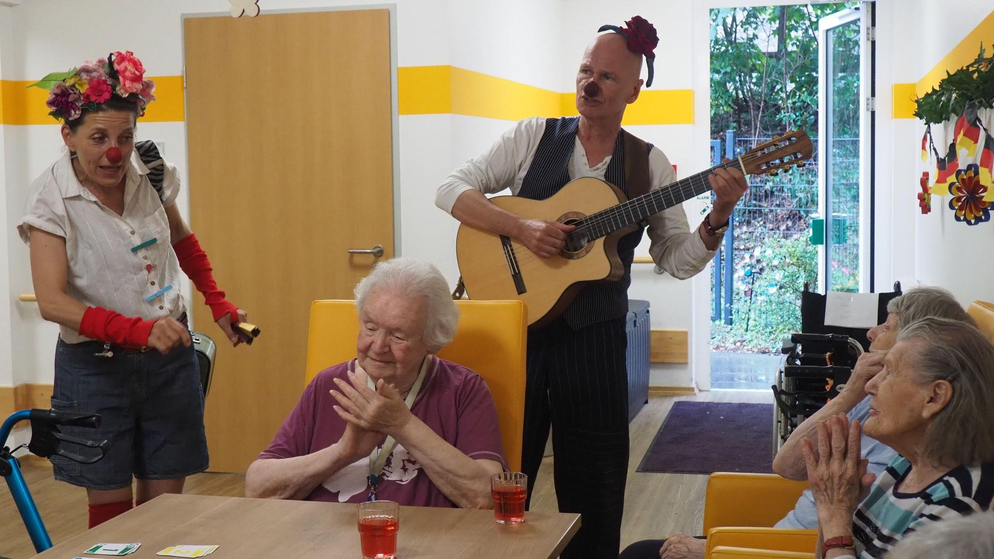 Ein Clown spielt Gitarre, ein weiblicher Clown links im Bild singt für die Seniorinnen, die begeistert klatschen.