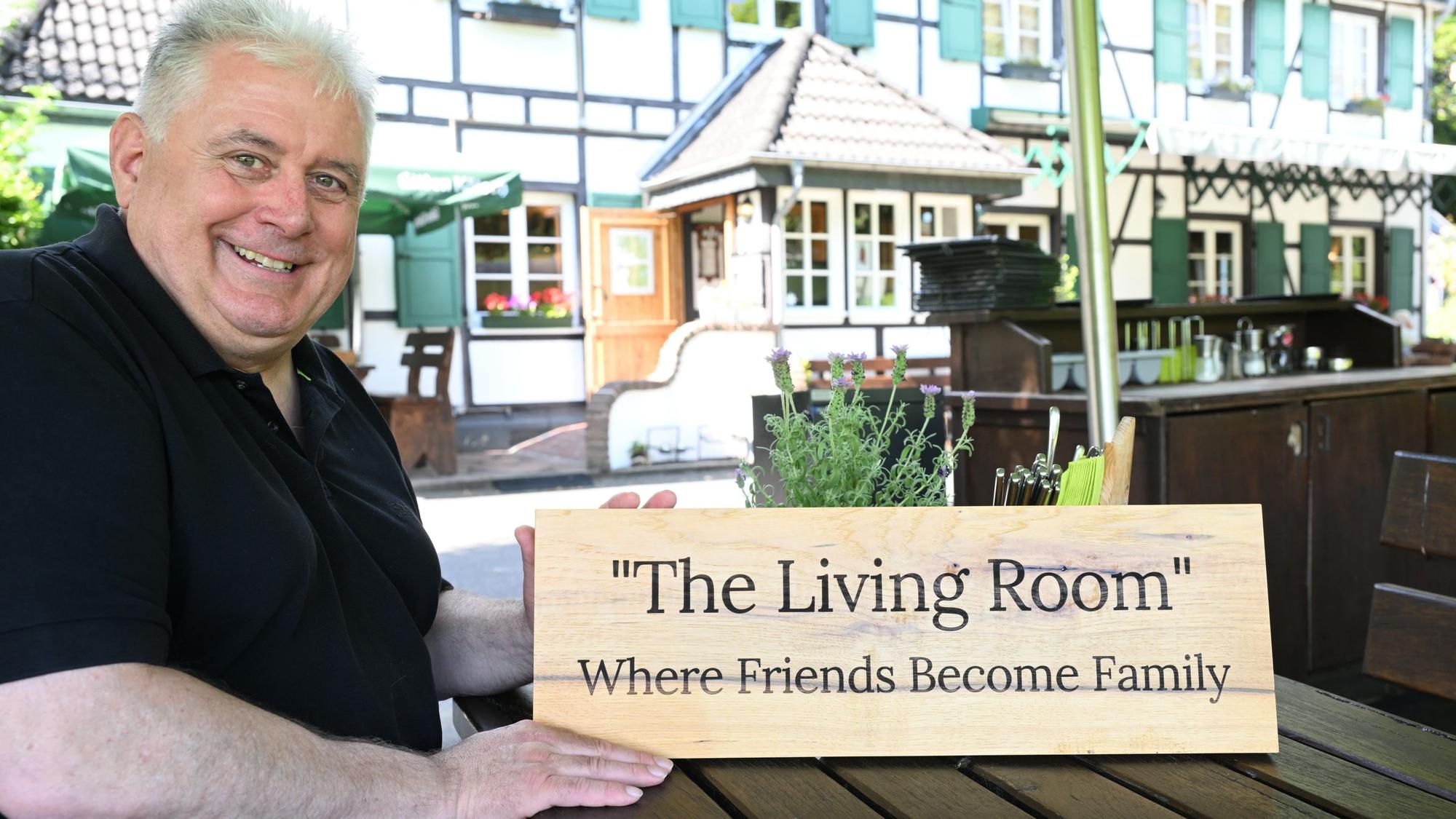 Ein Mann sitzt vor einem bergischen Fachwerklokal und hält ein Holzschild mit der Aufschrift: „The Living Room. Where Friends Become Family“ in der Hand.